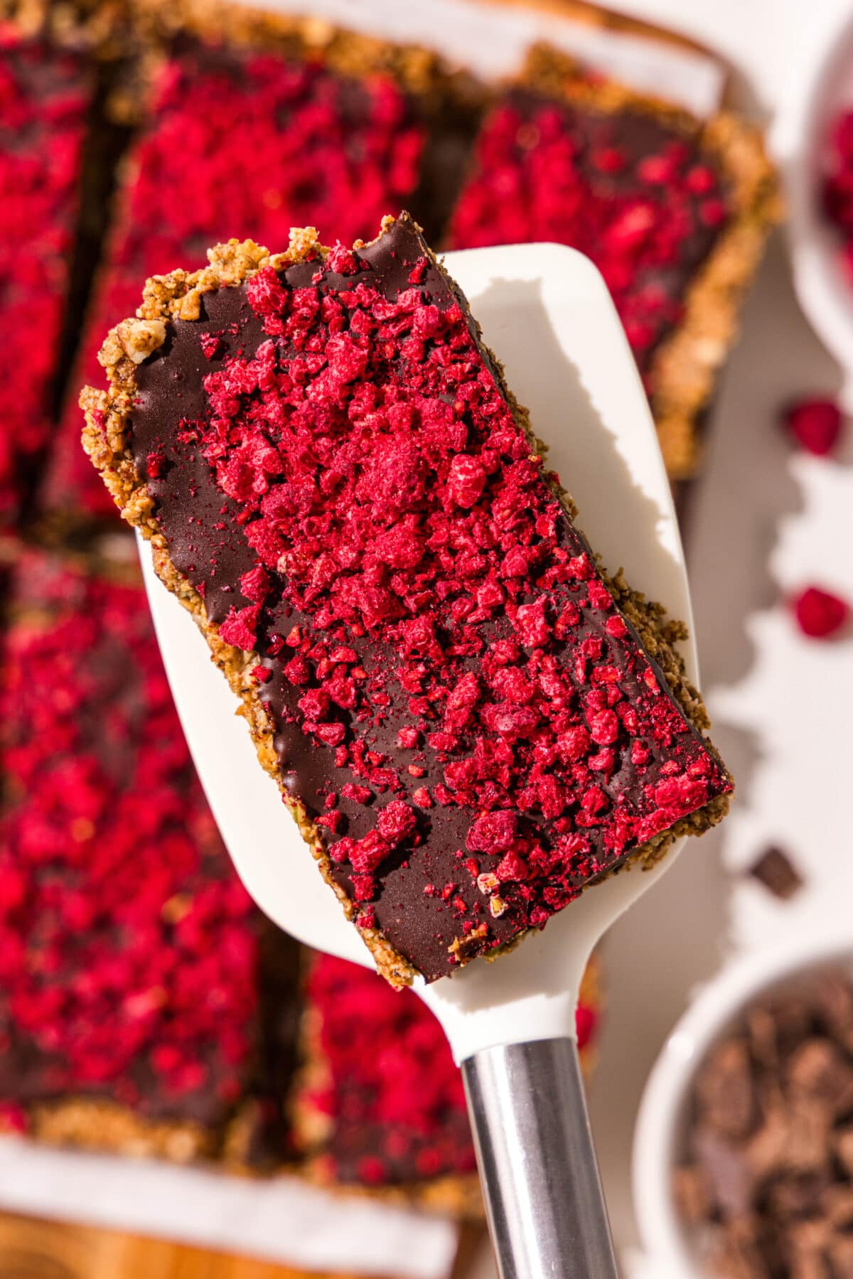 One dark chocolate raspberry cereal bar on a white rubber spatula over the remaining cereal bars on a wooden cutting board.