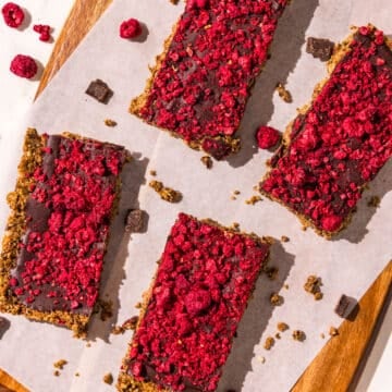 Four cereal bars topped with dark chocolate and freeze dried raspberries on a cutting board covered with a piece of parchment paper. Around the board is freeze dried raspberries and chocolate chips.