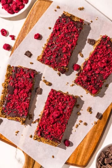 Four cereal bars topped with dark chocolate and freeze dried raspberries on a cutting board covered with a piece of parchment paper. Around the board is freeze dried raspberries and chocolate chips.