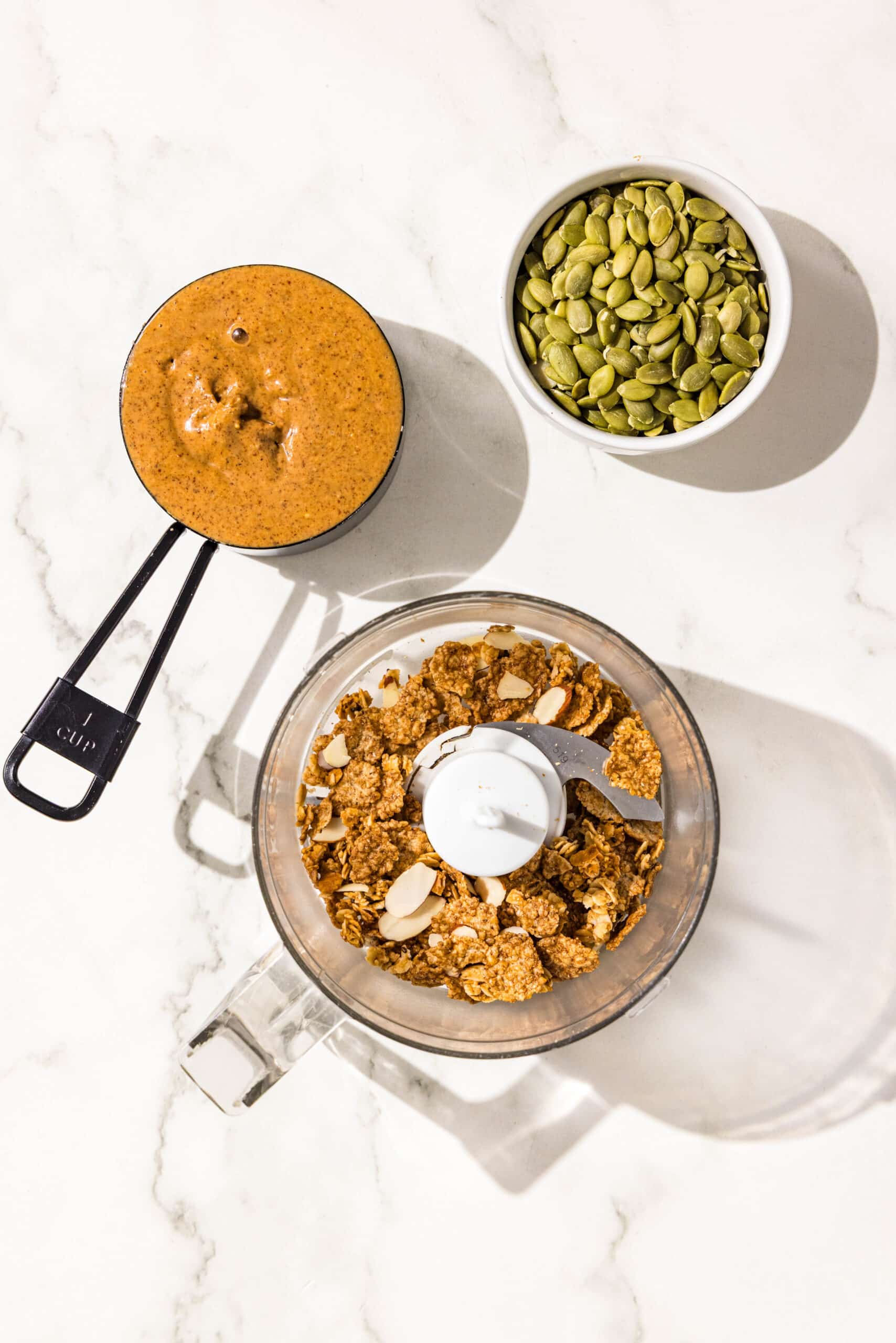 A small food processor filled with healthy cereal. Beside the food processor is a black 1 cup measuring cup filled with almond butter and a bowl of pumpkin seeds.