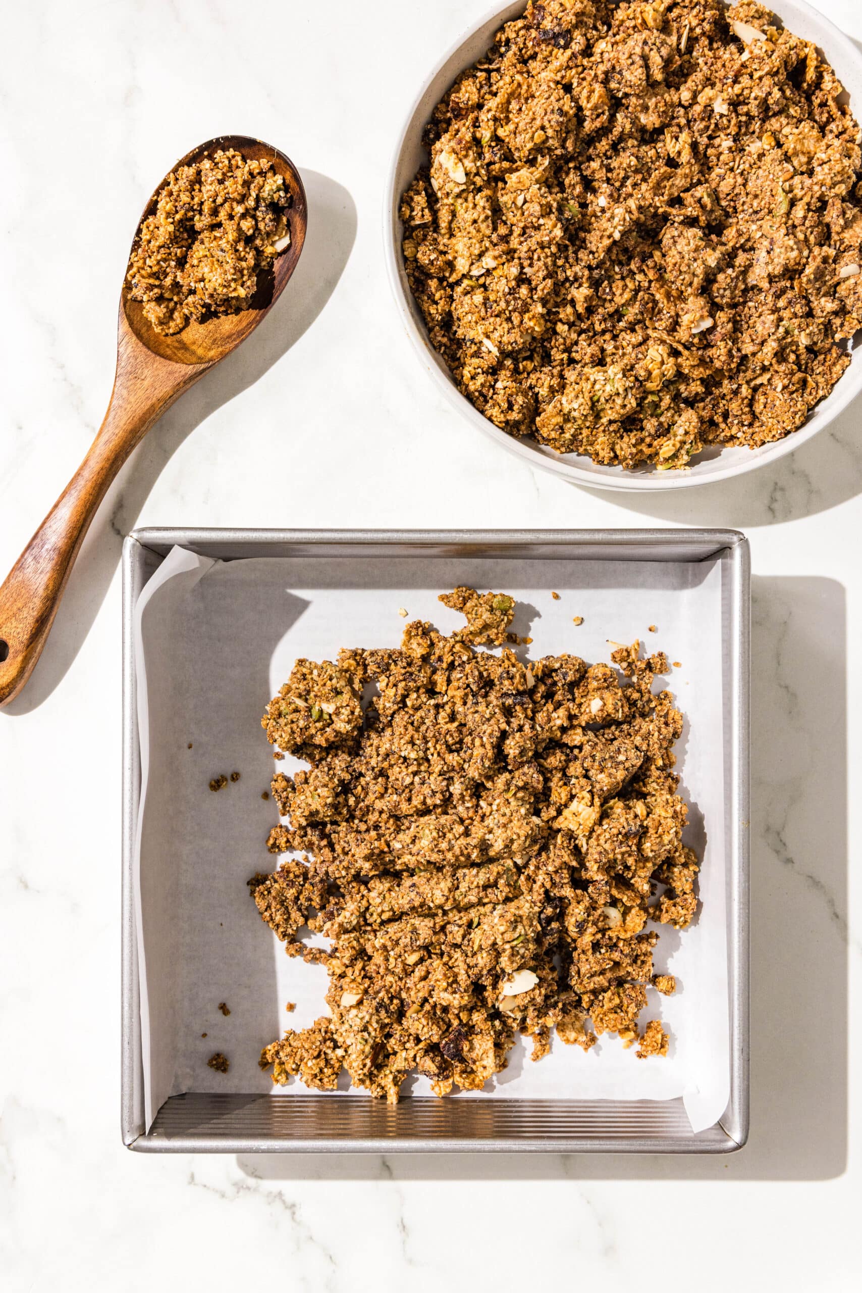 A 9x9 baking pan lined with parchment paper with a scoop of cereal bar dough in it. Beside the pan is a wooden spoon with some dough on it and a white bowl filled with the remaining cereal bar dough.