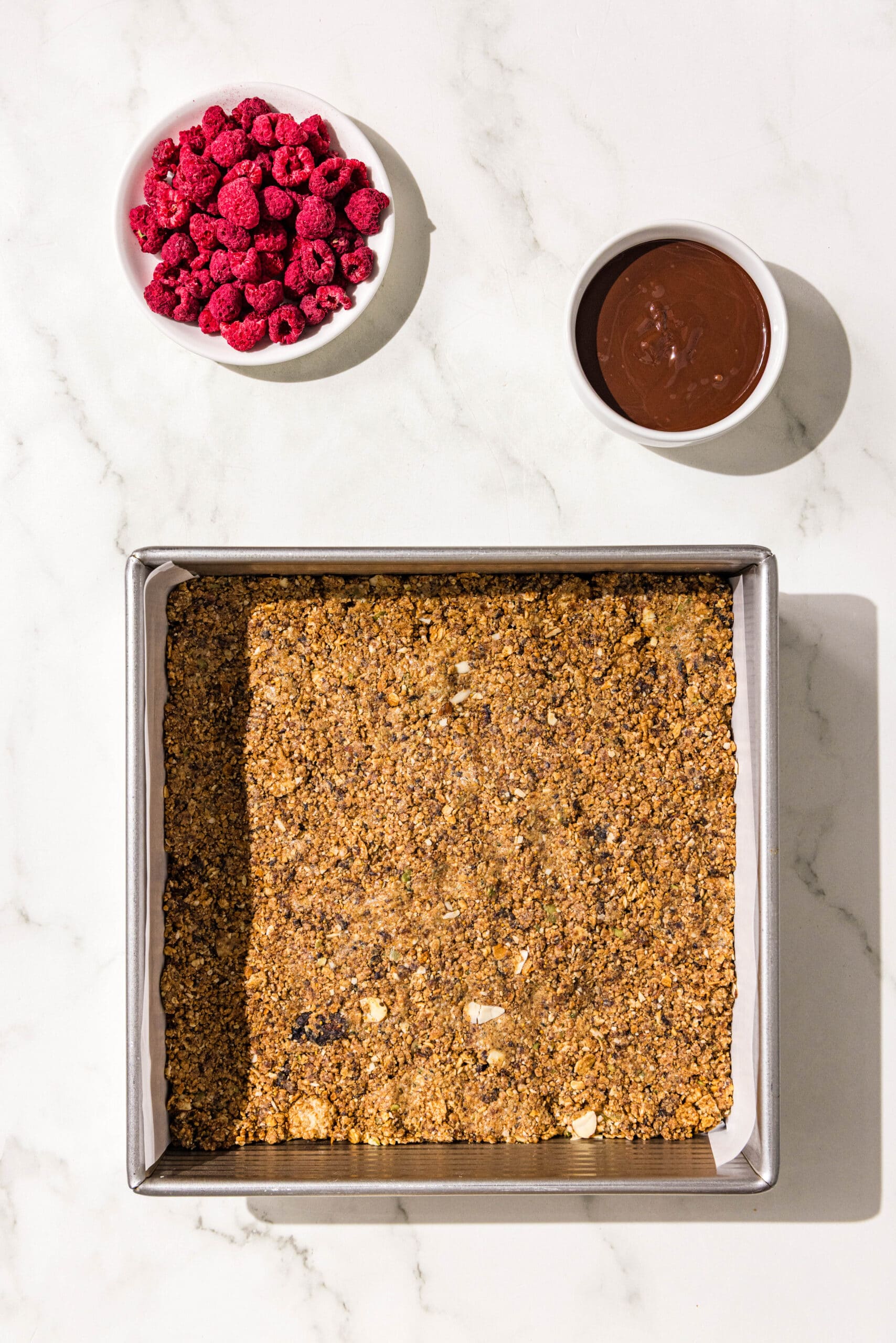 A 9x9 baking pan lined with parchment paper with cereal bar dough pressed into it. Above the pan is a small dish of freeze dried raspberries and bowl of melted dark chocolate.