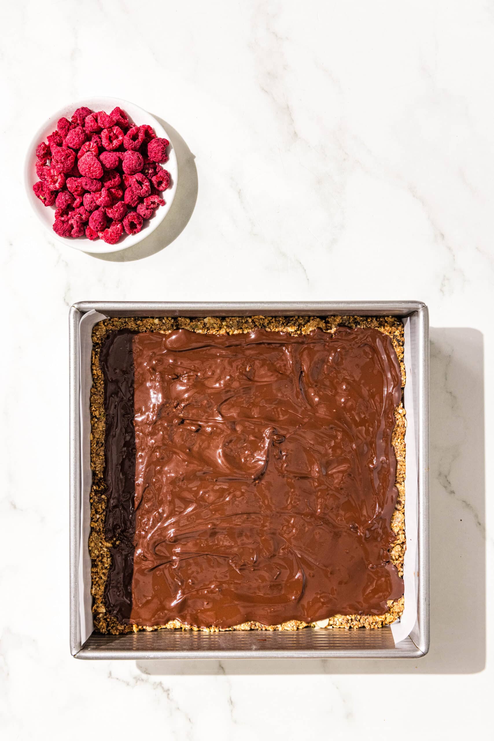 A 9x9 baking pan lined with parchment paper filled with cereal bars coated with melted dark chocolate on top. Beside the pan is a small dish of freeze dried raspberries.