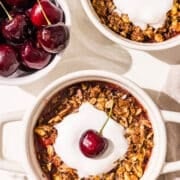 Two mini white ramekin dishes with cherry cobbler in them and a dollop of melted whipped cream in the center. The bottom one has a fresh cherry in the whipped cream. Beside the dishes is a small bowl filled with fresh cherries.