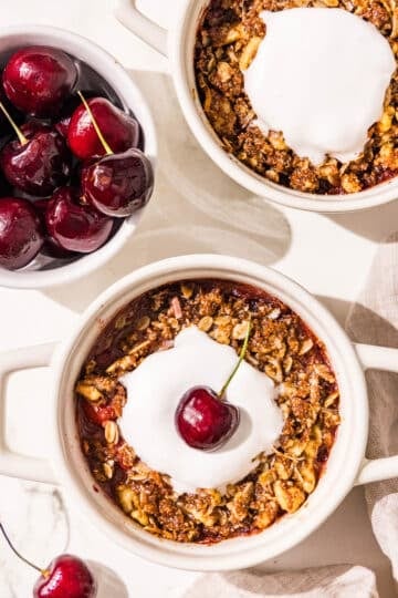 Two mini white ramekin dishes with cherry cobbler in them and a dollop of melted whipped cream in the center. The bottom one has a fresh cherry in the whipped cream. Beside the dishes is a small bowl filled with fresh cherries.