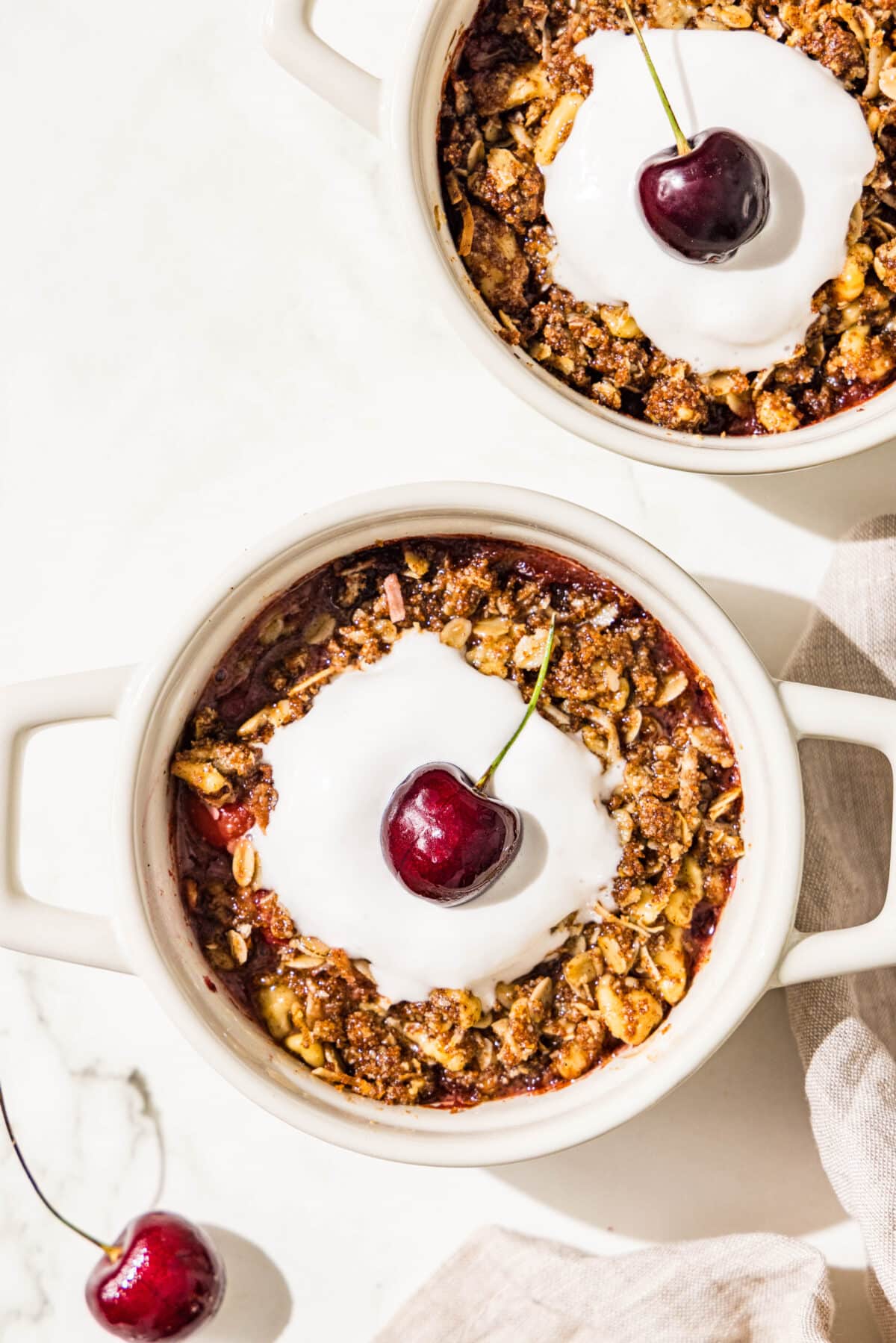 Two mini white ramekin dishes with cherry cobbler, whipped cream and a cherry in the center. Beside the dishes is a cherry on the counter and a linen towel.