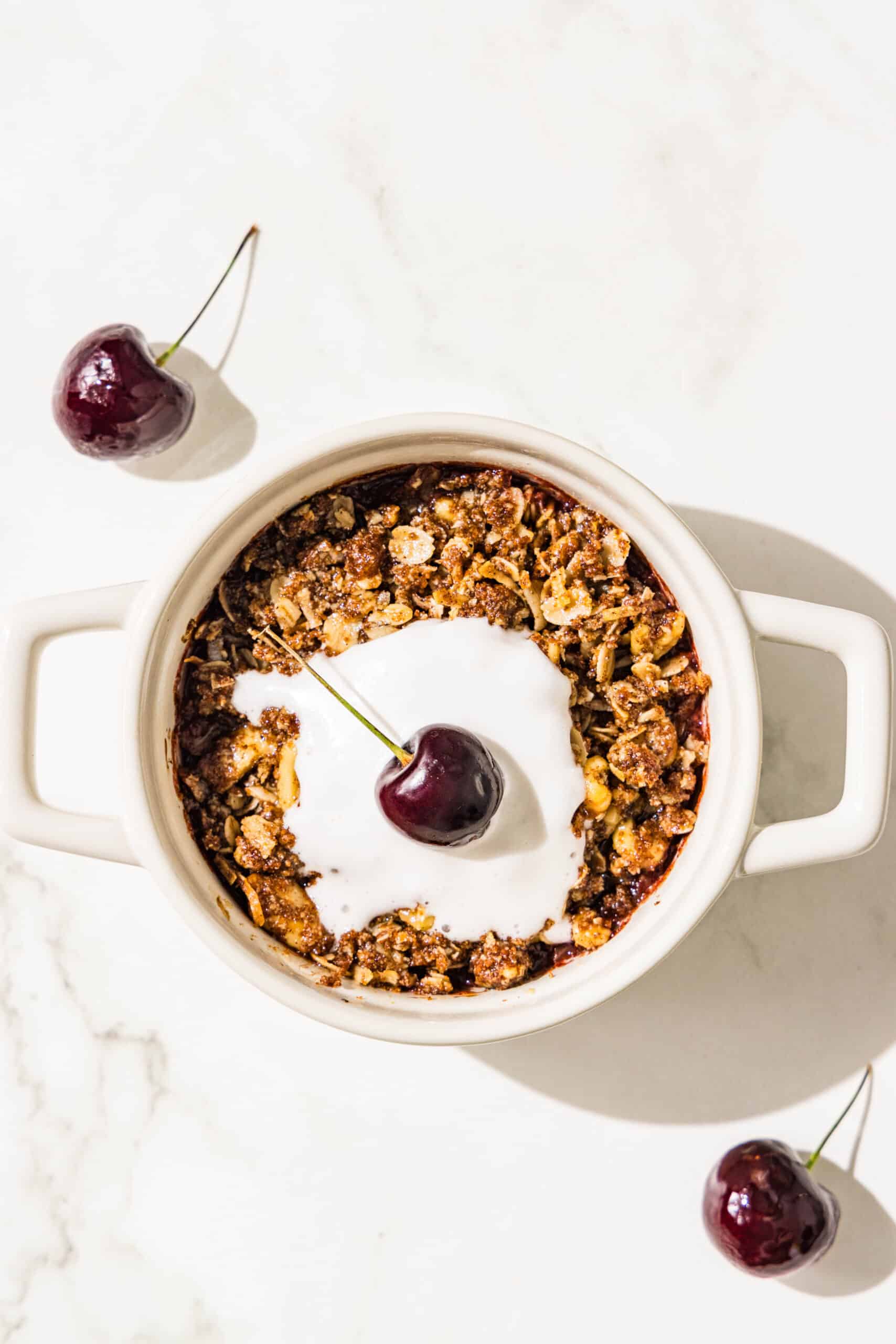 A white mini ramekin dish filled with a fruit crumble and melted whipped cream in the center. Beside the dish on the counter is a cherry.