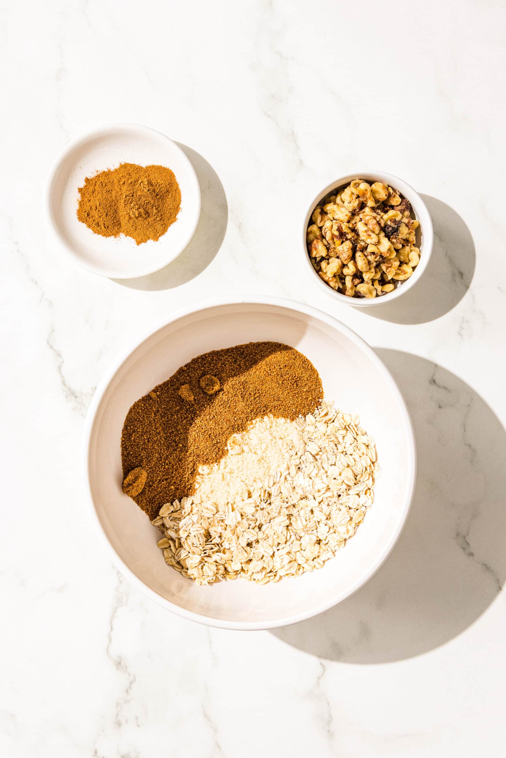 A large white bowl with coconut sugar, coconut, and oats in it. Beside the bowl is two small bowls filled with walnuts and cinnamon.