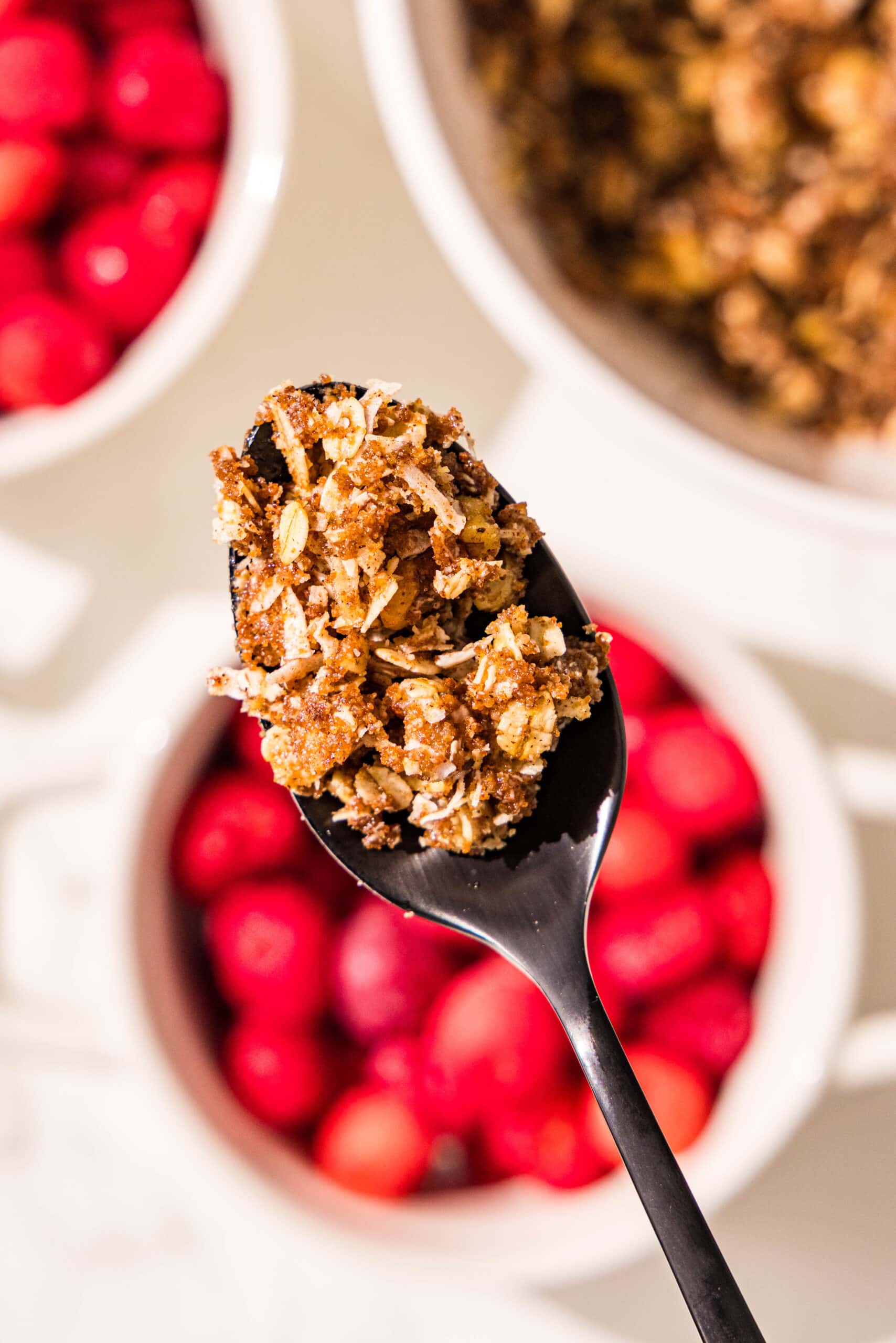 Two white ramekin dishes filled with frozen cherries. Beside those is a large bowl filled with oat crumble topping. A black spoon is in the center of the image in focus holding a bite of the crumble topping.