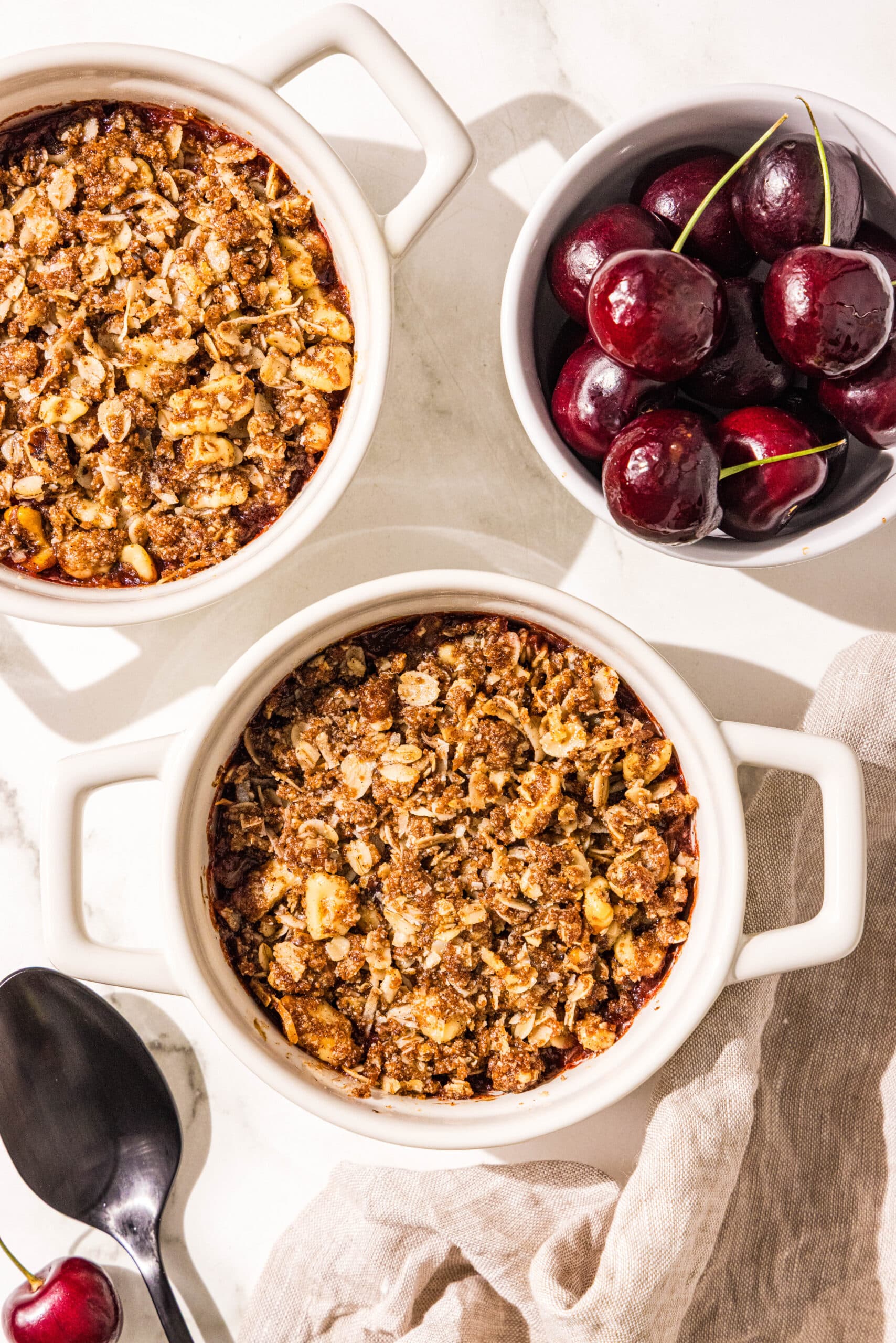 Two white ramekin dishes filled with cherries topped with oat crumble. Beside those is a small bowl of fresh cherries, a brown linen towel, and a black spoon.