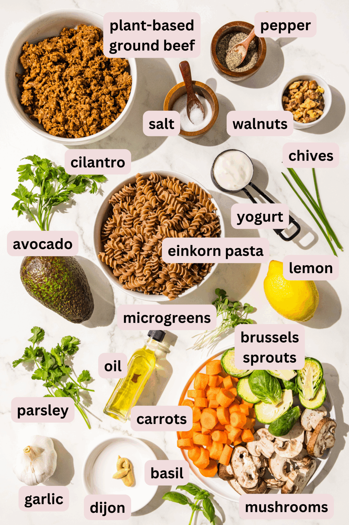 Ingredients needed to make hormone healthy pasta bowls laid out on the counter and labeled including: plant based ground beef, pepper, salt, walnuts, cilantro, chives, yogurt, avocado, einkorn pasta, lemon, parsley, carrots, oil, microgreens, brussels sprouts, mushrooms, basil, dijon, and garlic.