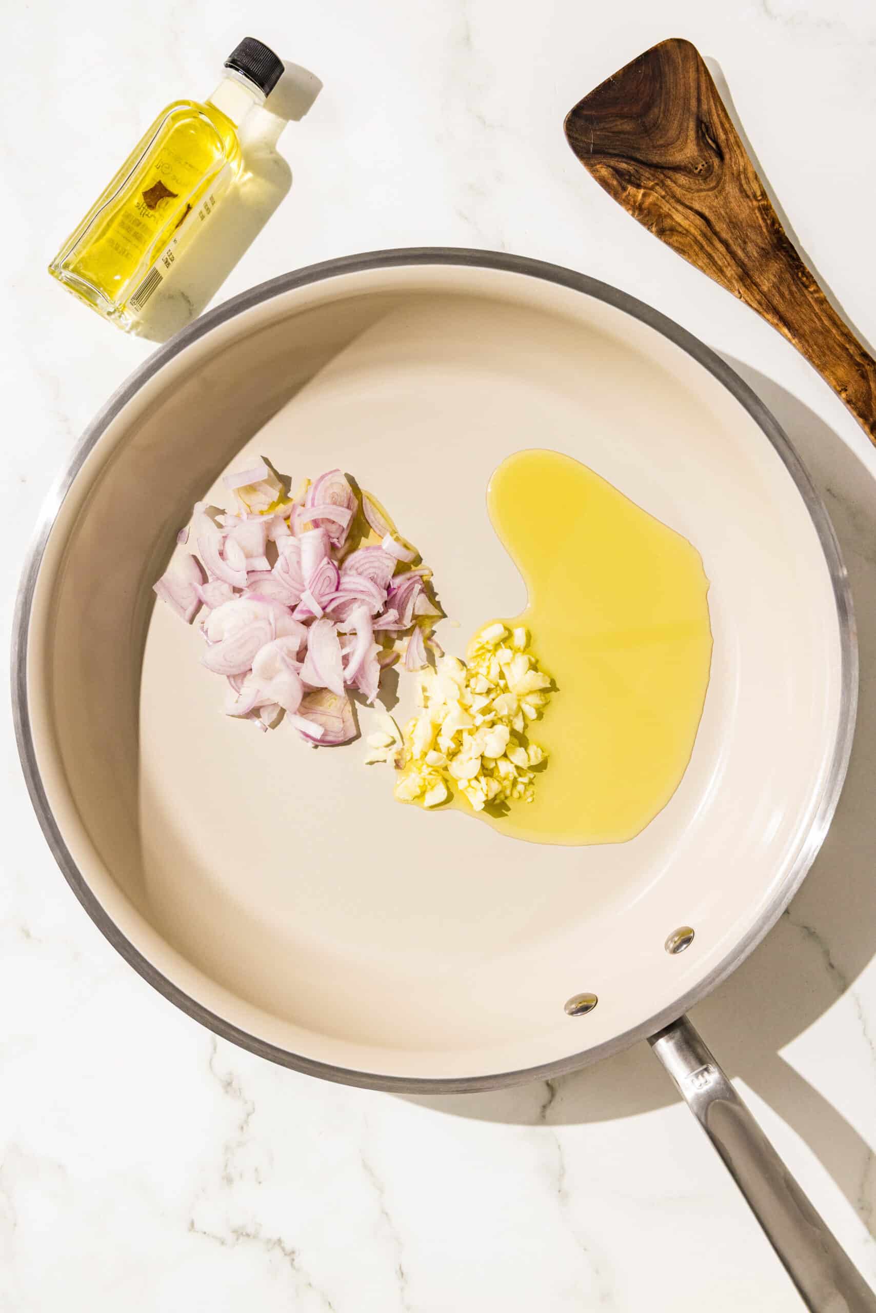 A white pan with oil, minced garlic and minced shallot in it. Beside the pan is a wooden spatula and a small bottle of oil.
