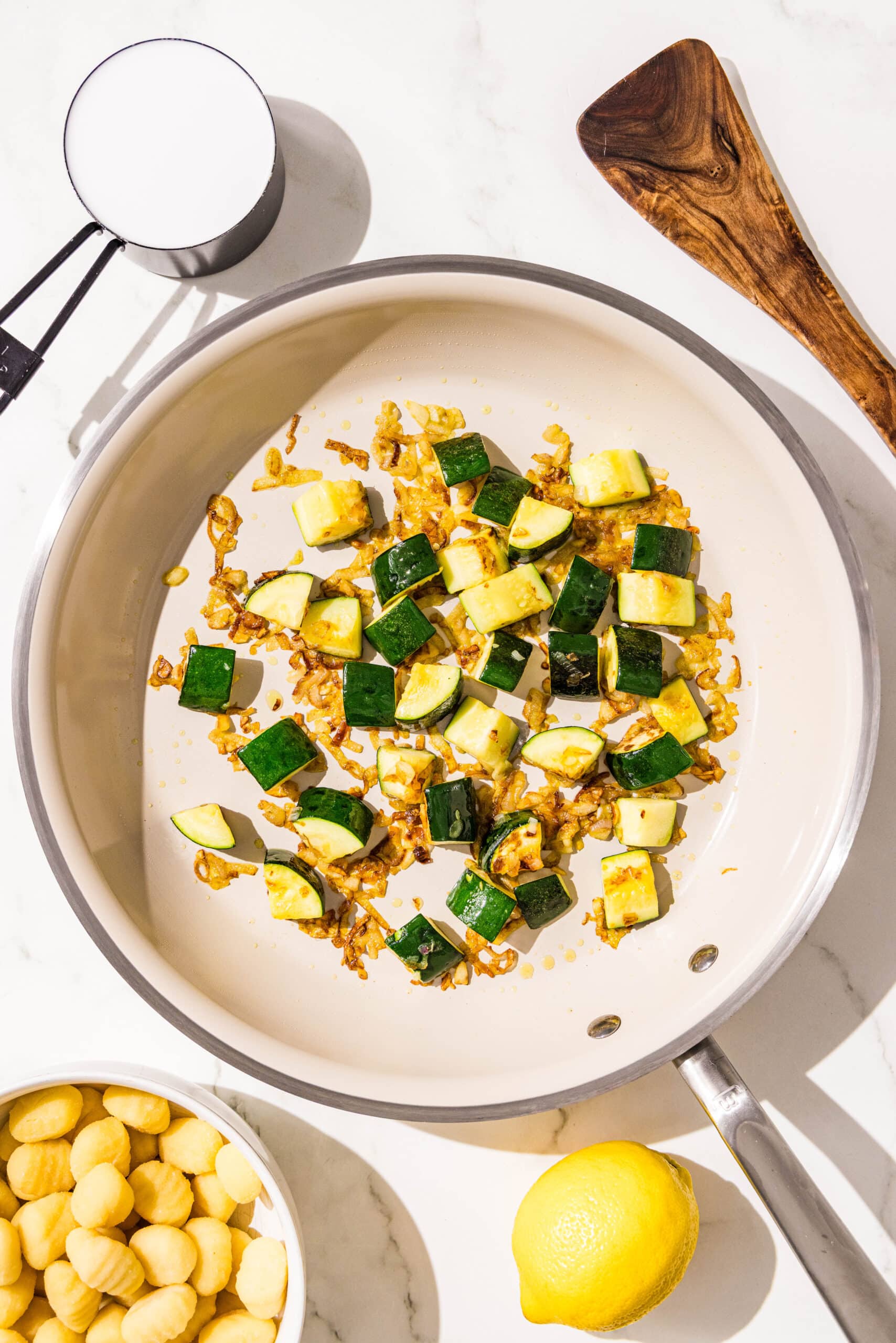 A pan filled with sauteed garlic and chopped zucchini. Around the pan on the counter is a whole lemon, a wooden spatula, a measuring cup filled with milk, and a small bowl gnocchi.
