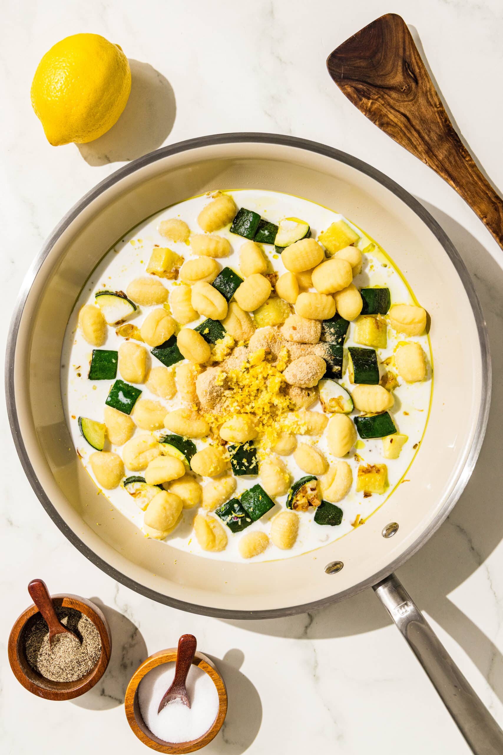 A pan filled with milk, zucchini, gnocchi, and garlic. Around the pan is a wooden spatula, a whole lemon, and two small wooden bowls of salt and pepper.