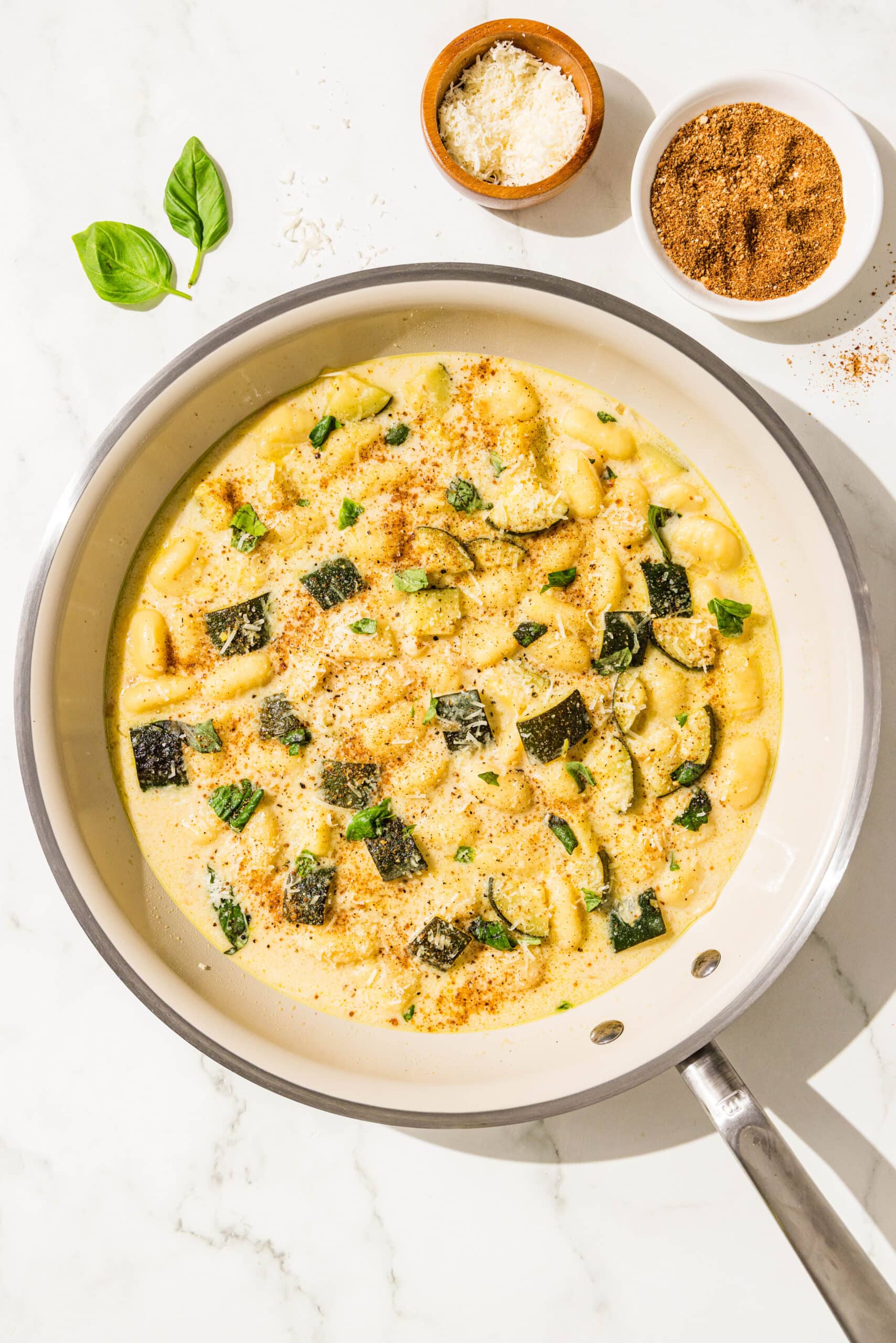 A pan filled with gnocchi, zucchini, and a yellow cream sauce. Around the pan is a small wooden bowl of parmesan and a white bowl of toasted breadcrumbs.