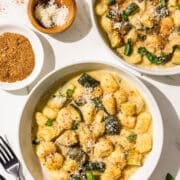 Two white bowls of a zuchinni and gnocchi dish topped with breadcrumbs and parmesan. Beside the bowls on the left is two black forks, a small bowl of breadcrumbs, a small bowl of parmesan and parmesan on the counter. On the right is two basil leaves on the counter.