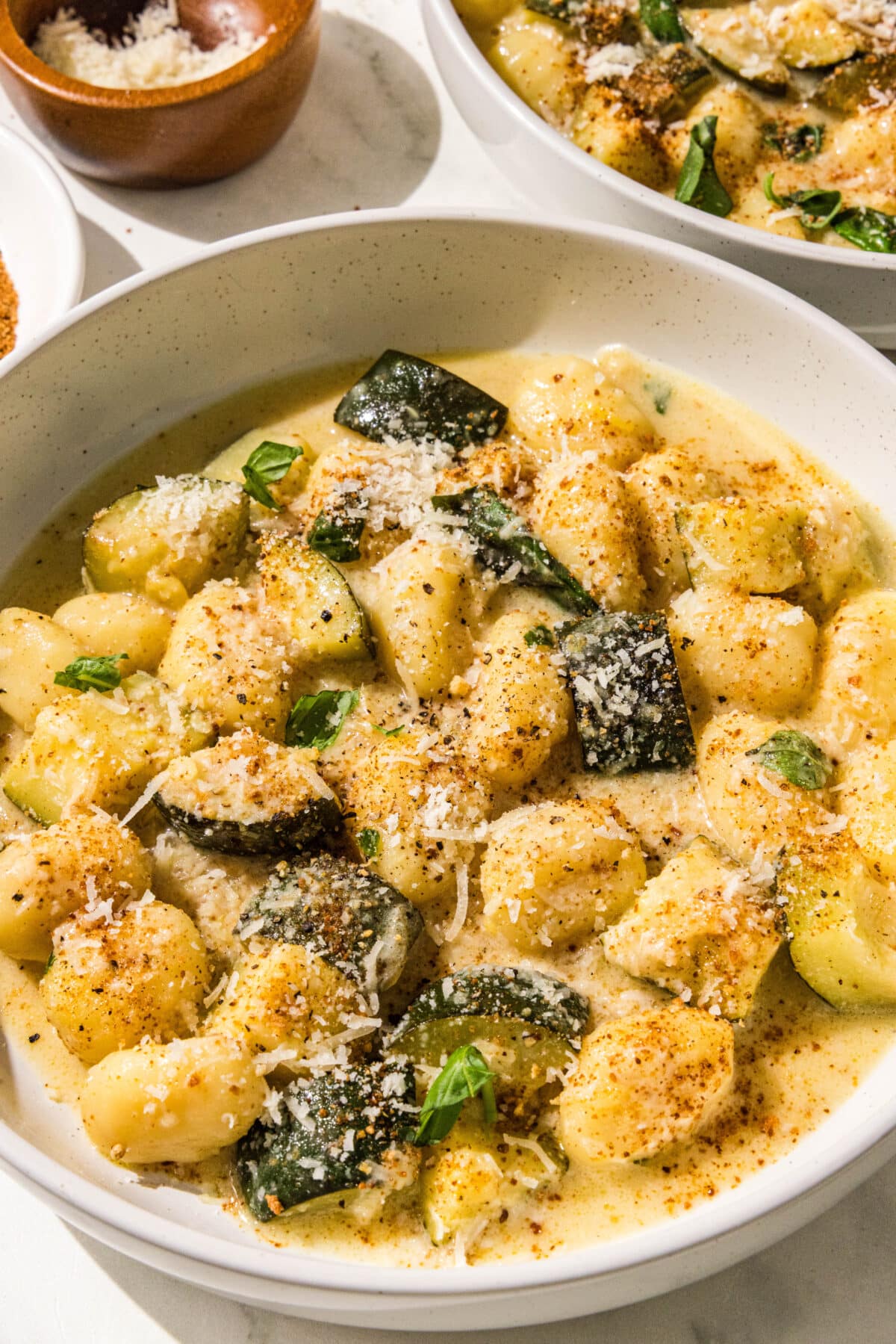 A white bowl filled with zuchinni, gnocchi, and a yellow cream sauce. In the background is another bowl and a small wooden bowl of parmesan shreds.