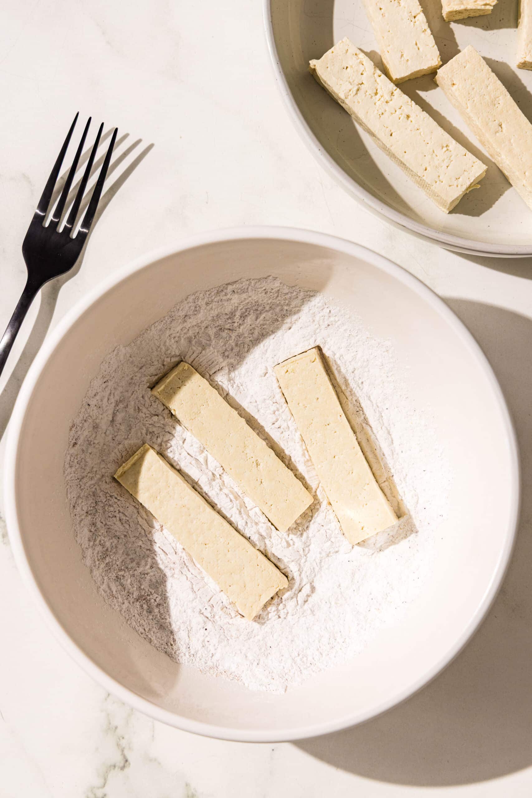 A white bowl filled with an arrowroot flour mixture with three sticks of tofu in it. Beside the bowl is a plate with remaining tofu and a black metal fork.