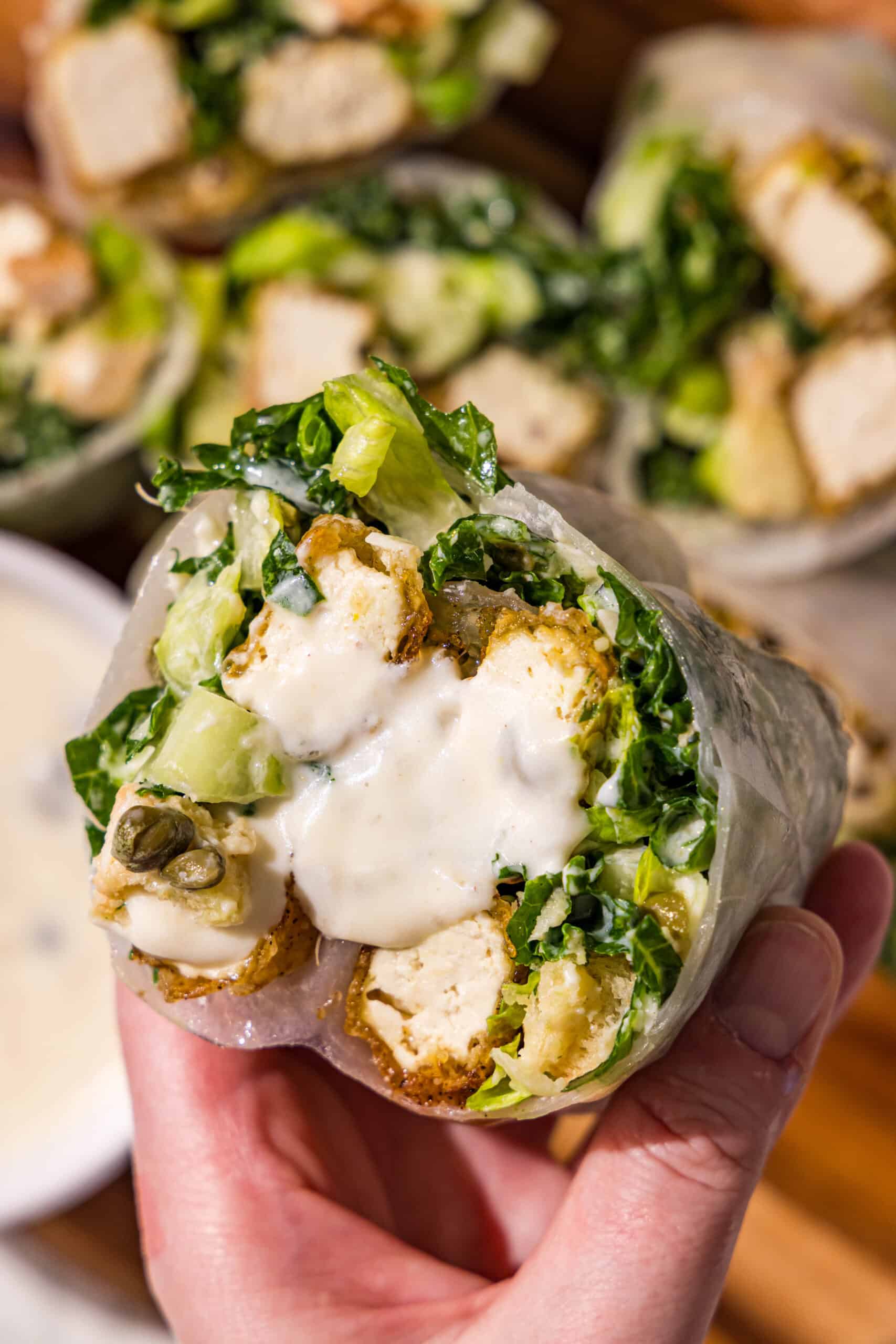 A caesar salad tofu spring roll being held by a hand and a small bowl of caesar dressing.