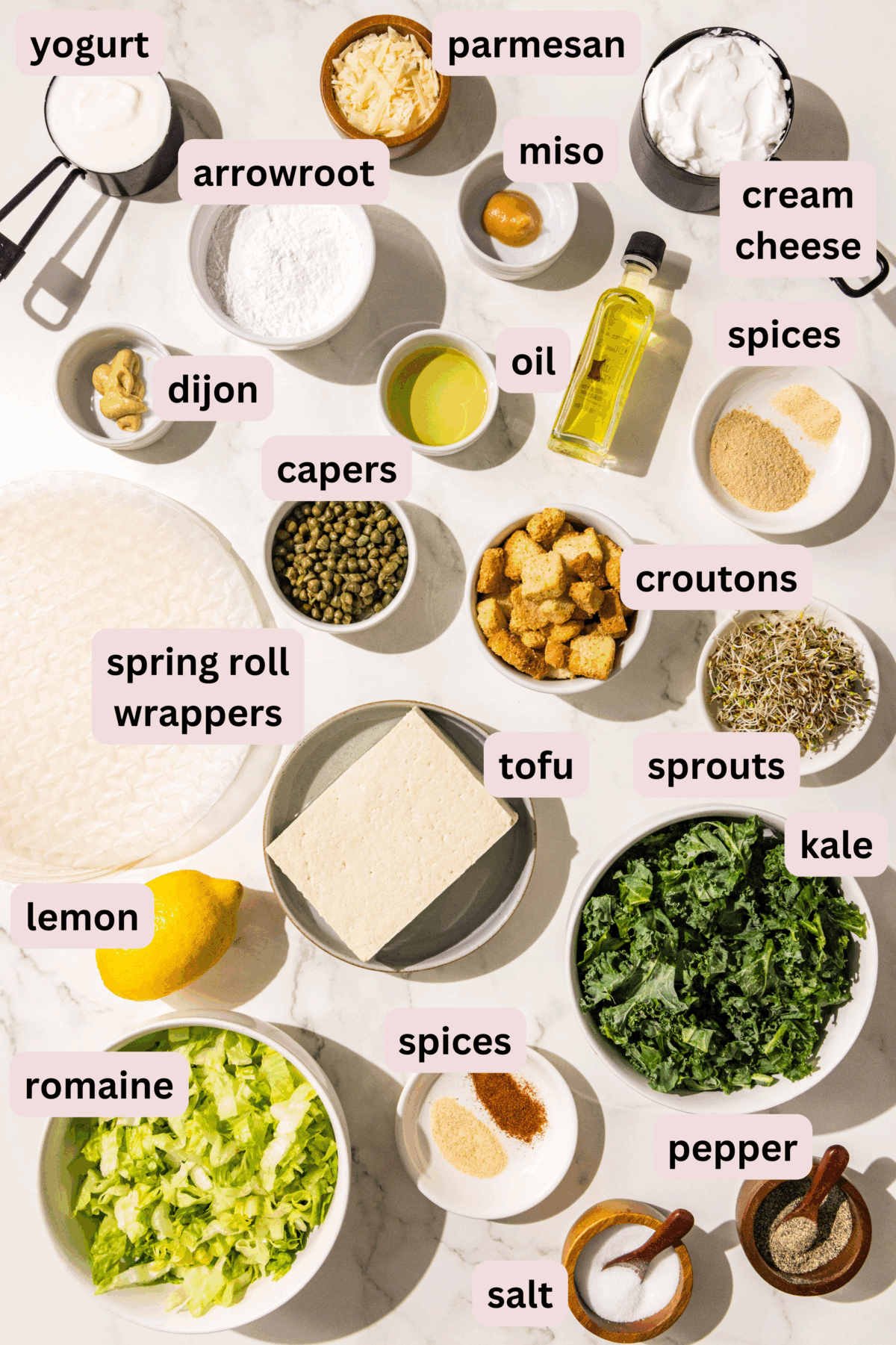 Ingredients laid out on a marble counter and labeled including yogurt, parmesan, cream cheese, miso, arrowroot powder, oil, dijon mustard, spices, capers, croutons, sprouts, spring roll wrappers, tofu, kale, romaine, a lemon, salt and pepper.