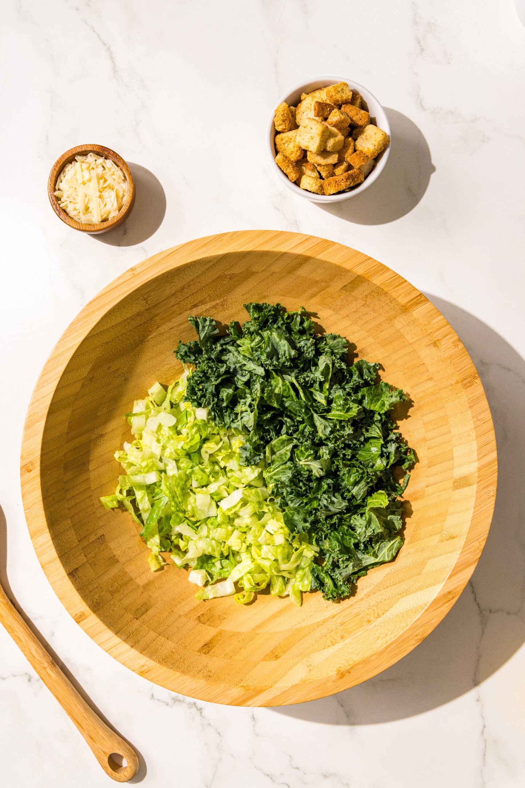 A large wooden bowl filled with half romaine and half kale. Beside the bowl is a wooden spoon and two small bowls of croutons and parmesan.