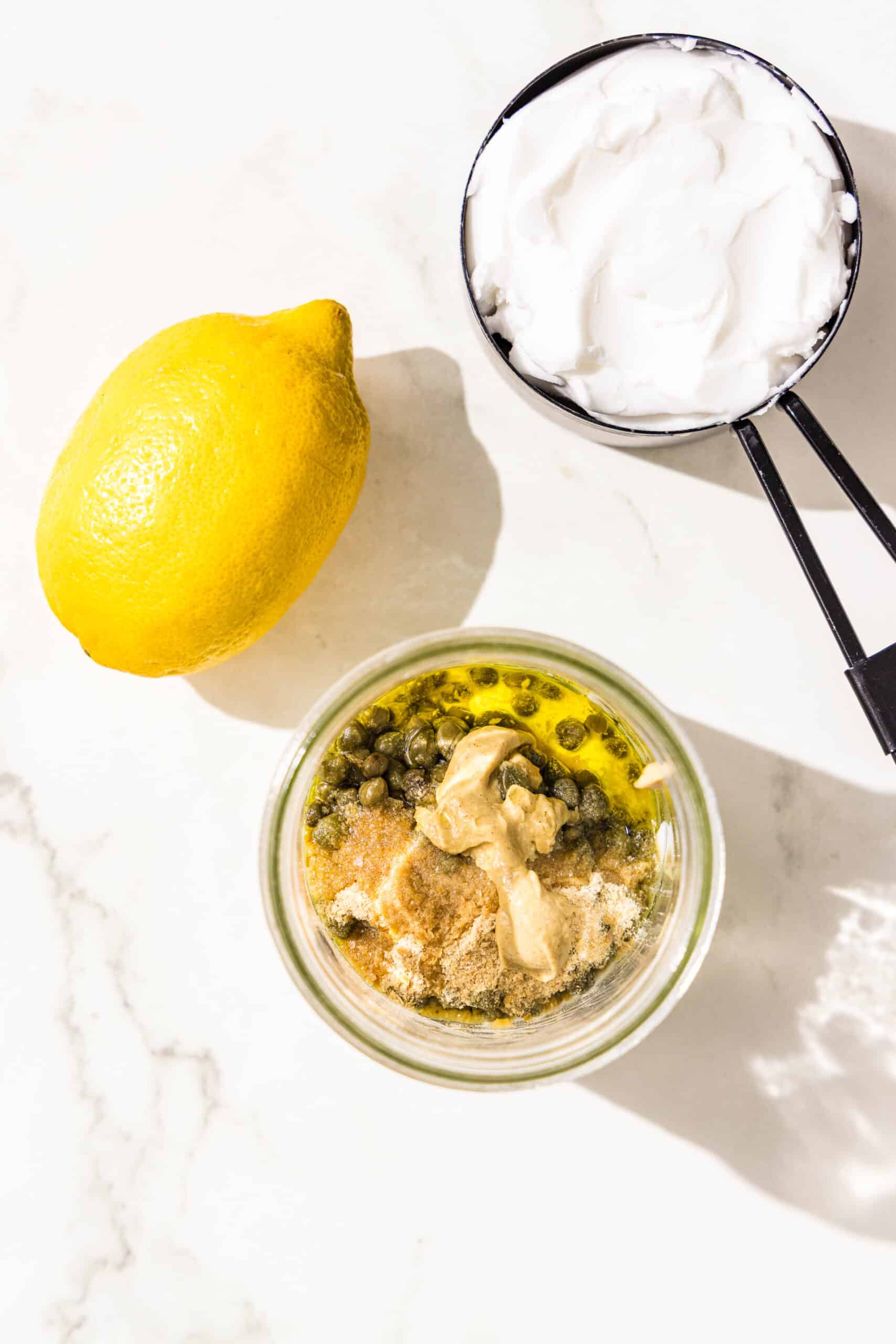 A glass jar of yogurt, dijon, capers, and oil. Beside the jar is a lemon and a black metal measuring cup filled with cream cheese.