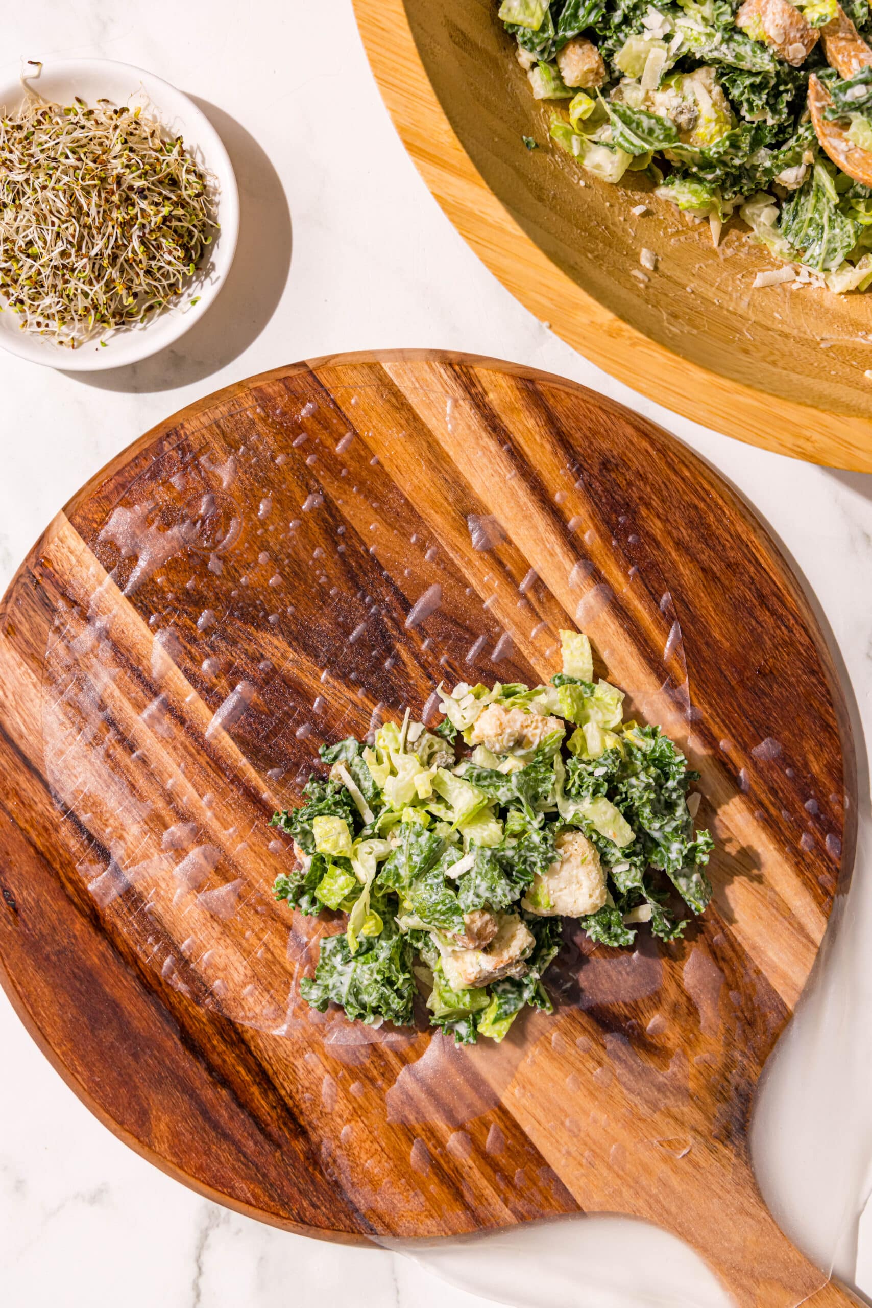 A wooden cutting board with a wet spring roll wrapper on it filled half with caesar salad. Beside the cutting board is a wooden bowl of salad and a small glass bowl of sprouts.