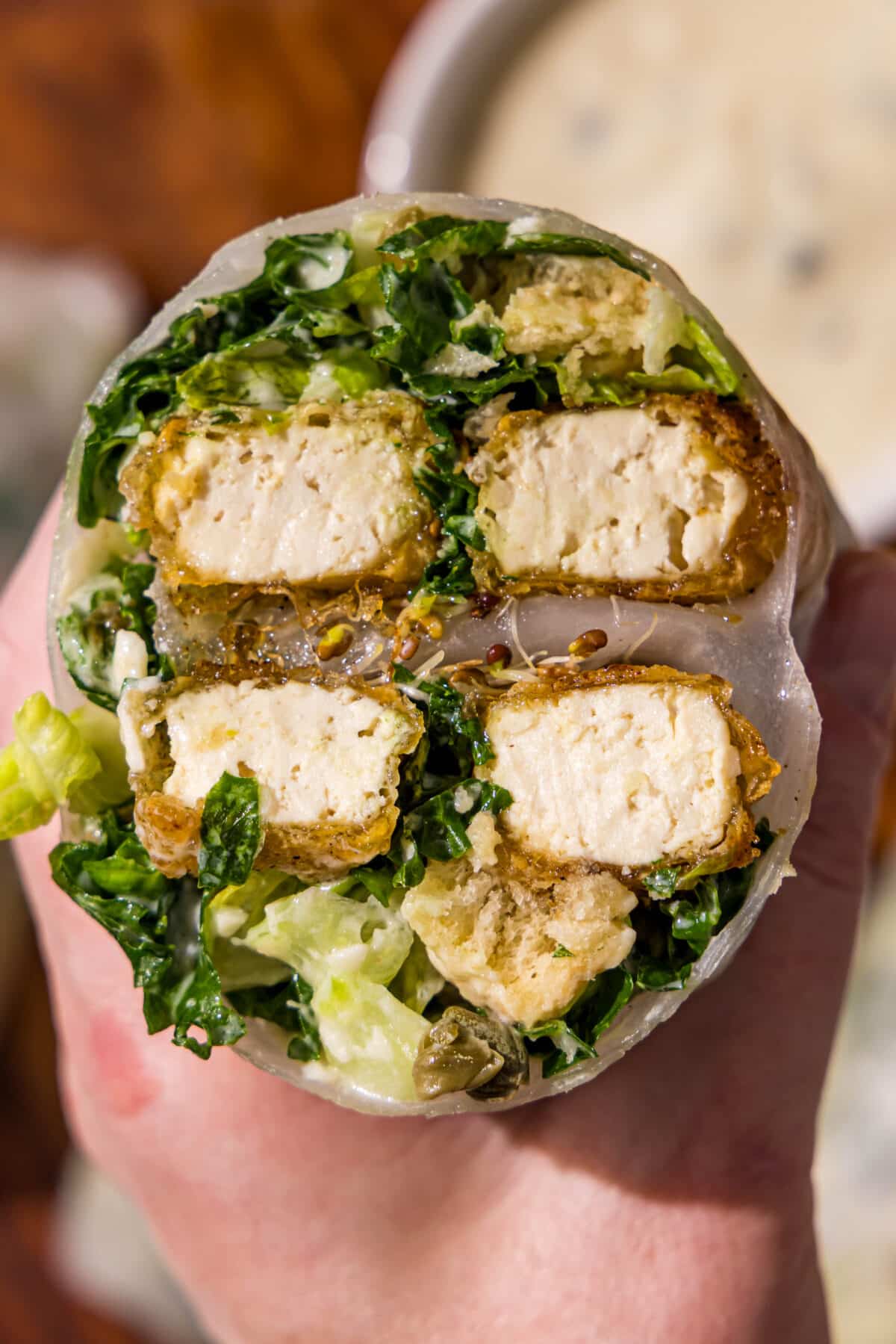 A caesar salad tofu spring roll cut in half held by a hand and a small bowl of caesar dressing on a wood cutting board.