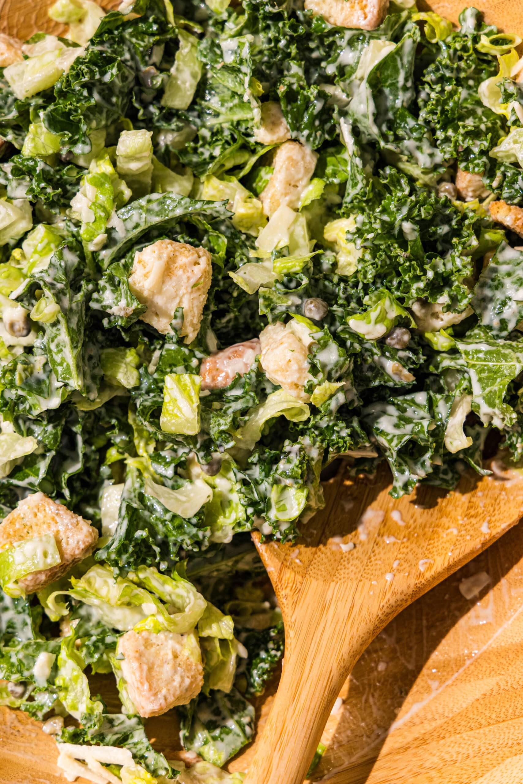 A wooden bowl filled with kale romaine caesar salad and a wooden spoon sticking out.