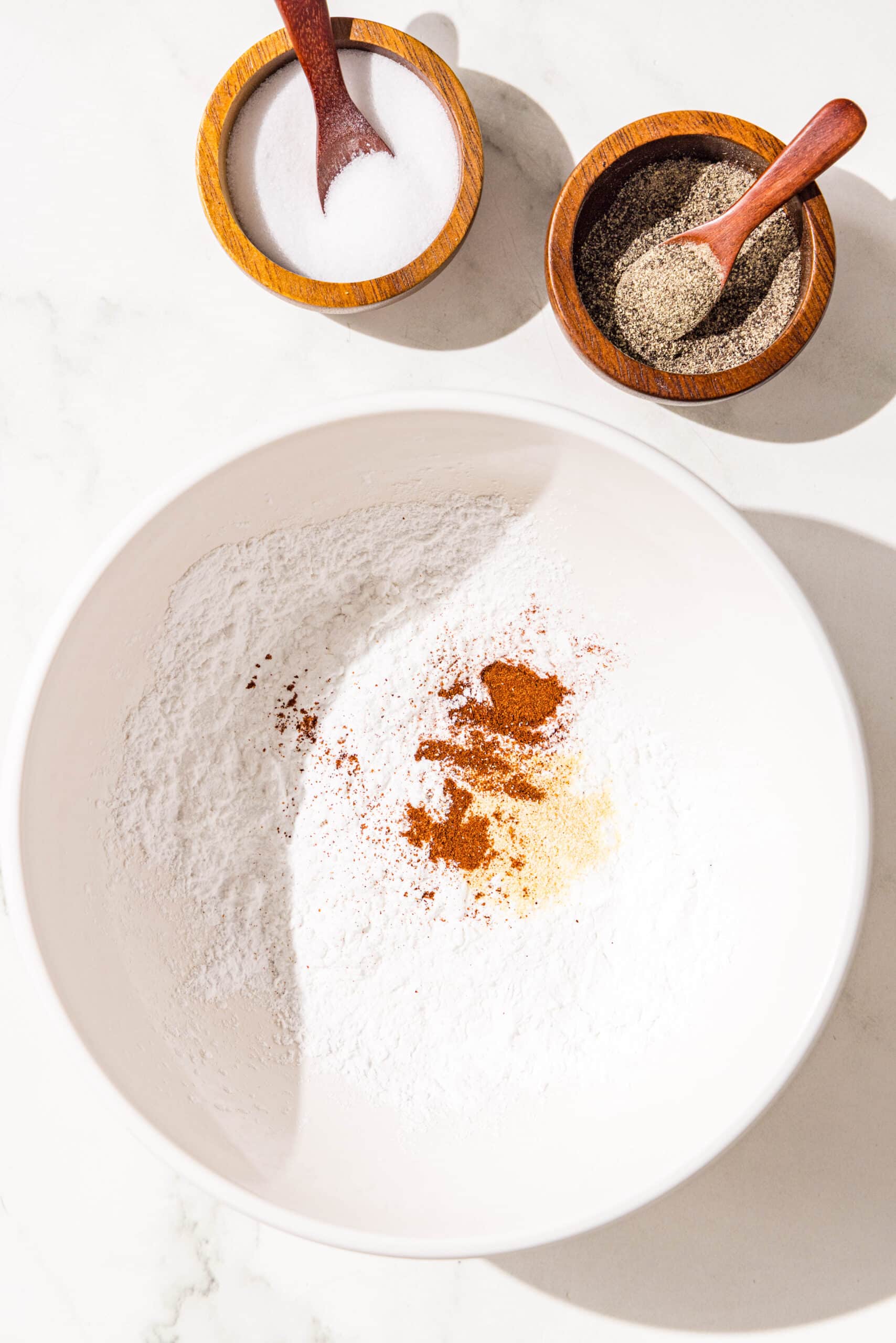A white glass bowl of arrowroot powder, garlic and paprika. Beside the bowl of two small wooden bowls of salt and pepper.