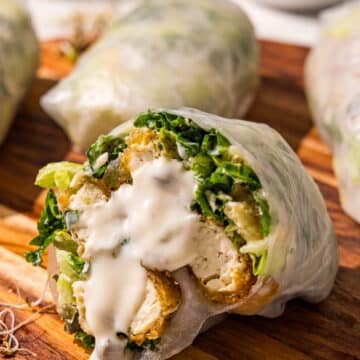 A wooden cutting board with three spring rolls on it and a fourth one cut in half with caesar dressing dripping down the center of it.