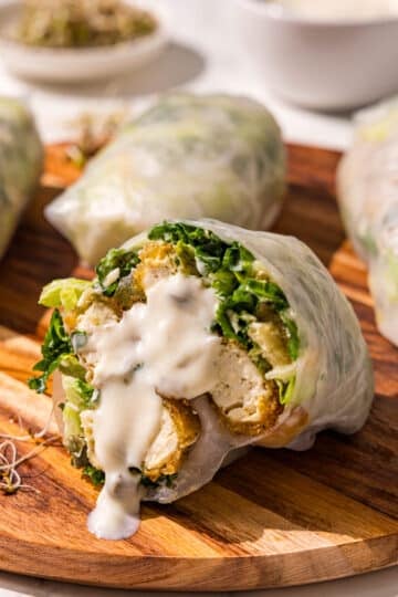 A wooden cutting board with three spring rolls on it and a fourth one cut in half with caesar dressing dripping down the center of it.