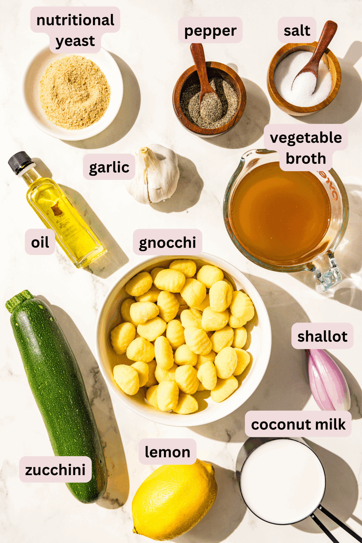 Ingredients laid out on a marble counter and labeled including: nutritional yeast, salt and pepper, garlic, vegetable broth, oil, gnocchi, shallot, zucchini, lemon, and coconut milk.