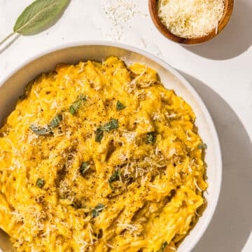 A bowl of pumpkin orzo garnished with fresh sage, black pepper, and olive oil. Beside the bowl is a small wooden bowl of parmesan and a fresh sage leaf.
