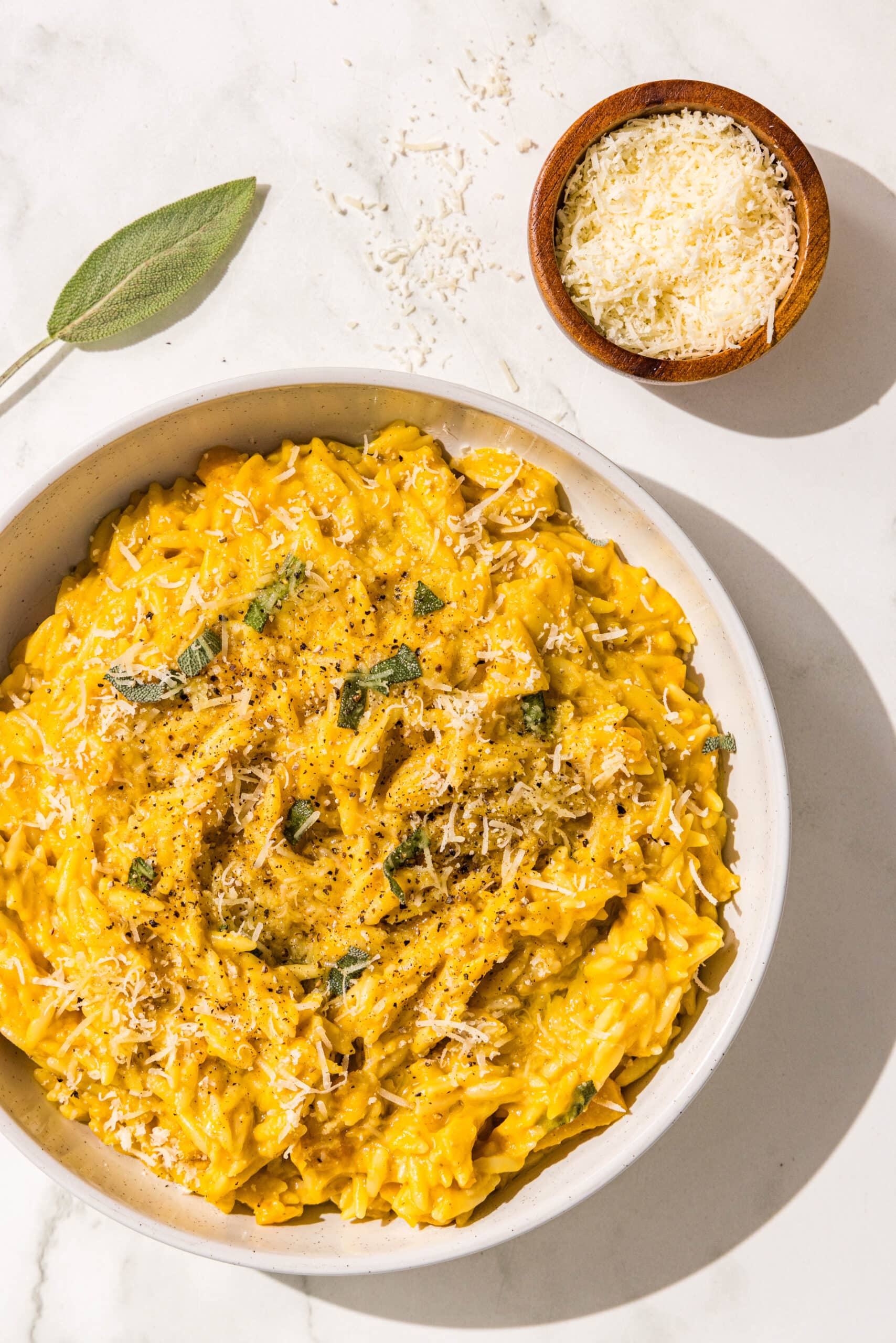 A bowl of pumpkin orzo garnished with fresh sage, black pepper, and olive oil. Beside the bowl is a small wooden bowl of parmesan and a fresh sage leaf.