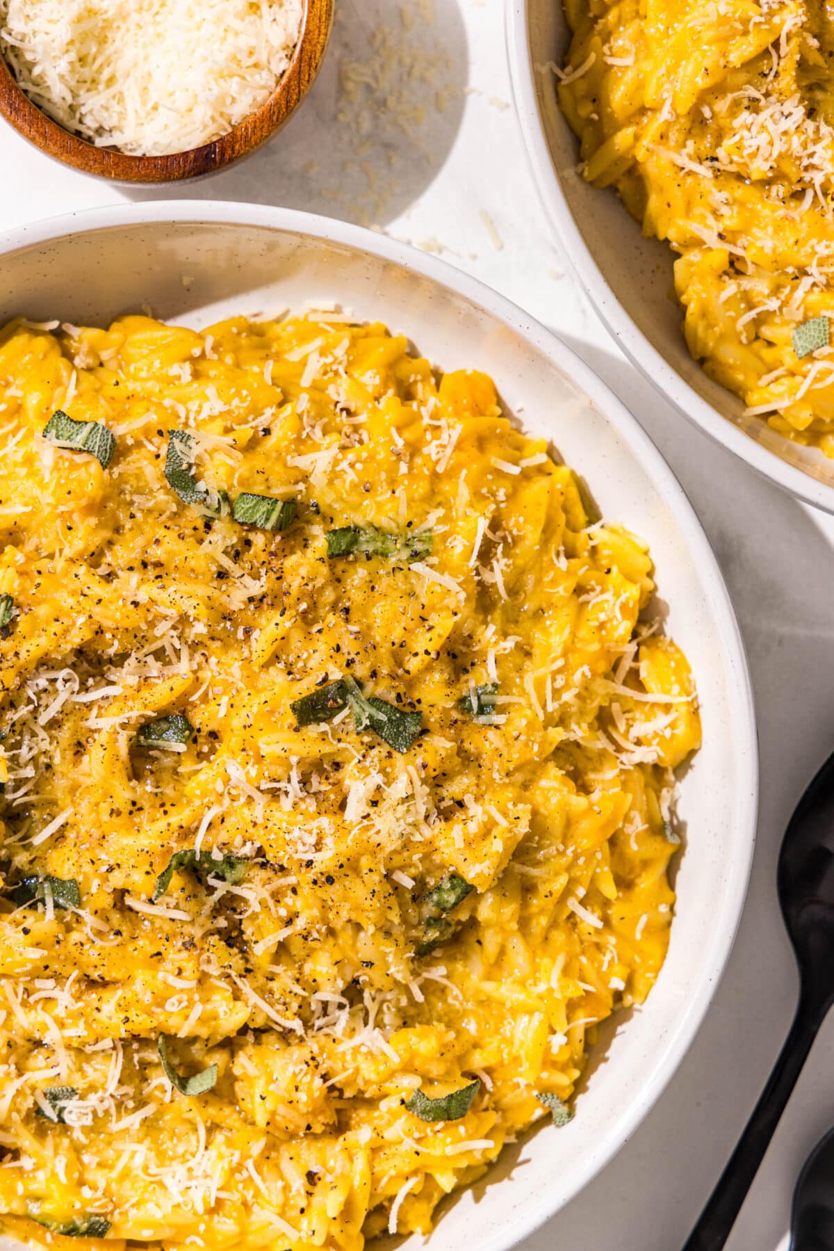 Two bowls of pumpkin orzo with two black spoons and a small wooden bowl of parmesan.
