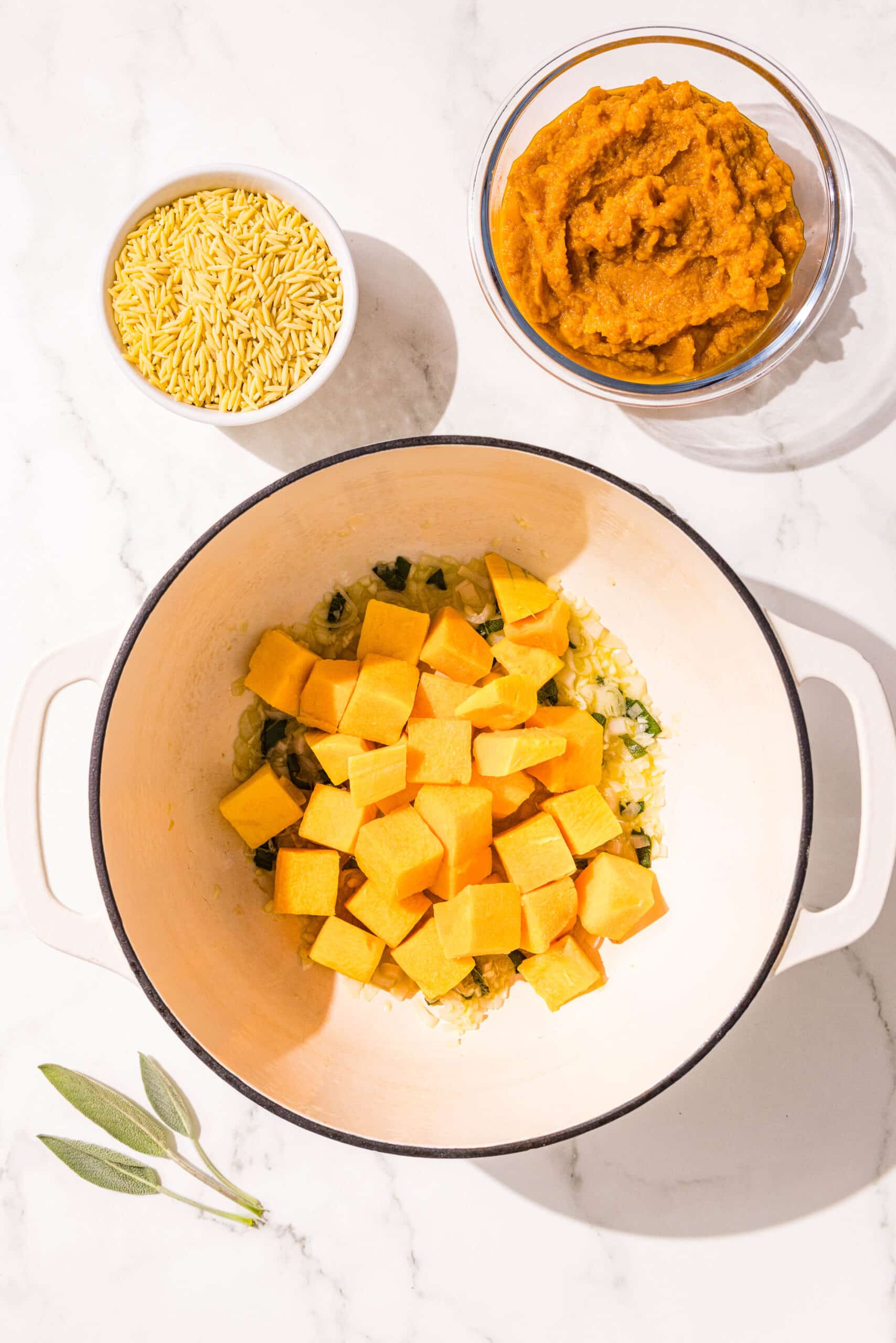 A large pot filled with oil, fresh sage, and fresh butternut squash. beside the pot is two small bowls of orzo and pumpkin puree.
