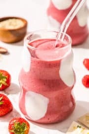 Two glasses of pink smoothie with a glass straw in it. Around the cups on the counter is fresh strawberries and a small wooden bowl of hemp seeds.