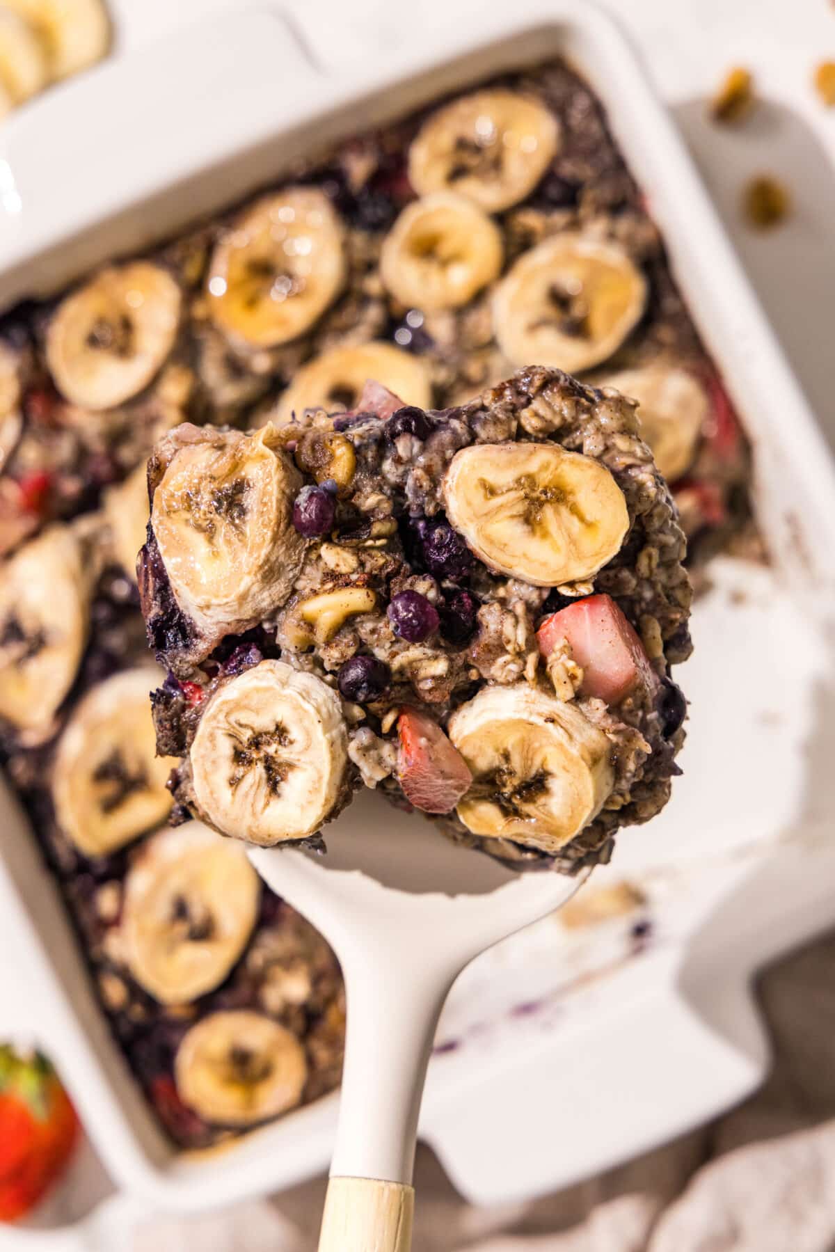 A 9x9 baking dish of berry baked oatmeal topped with sliced bananas and a spatula holding a slice of baked oatmeal on it.