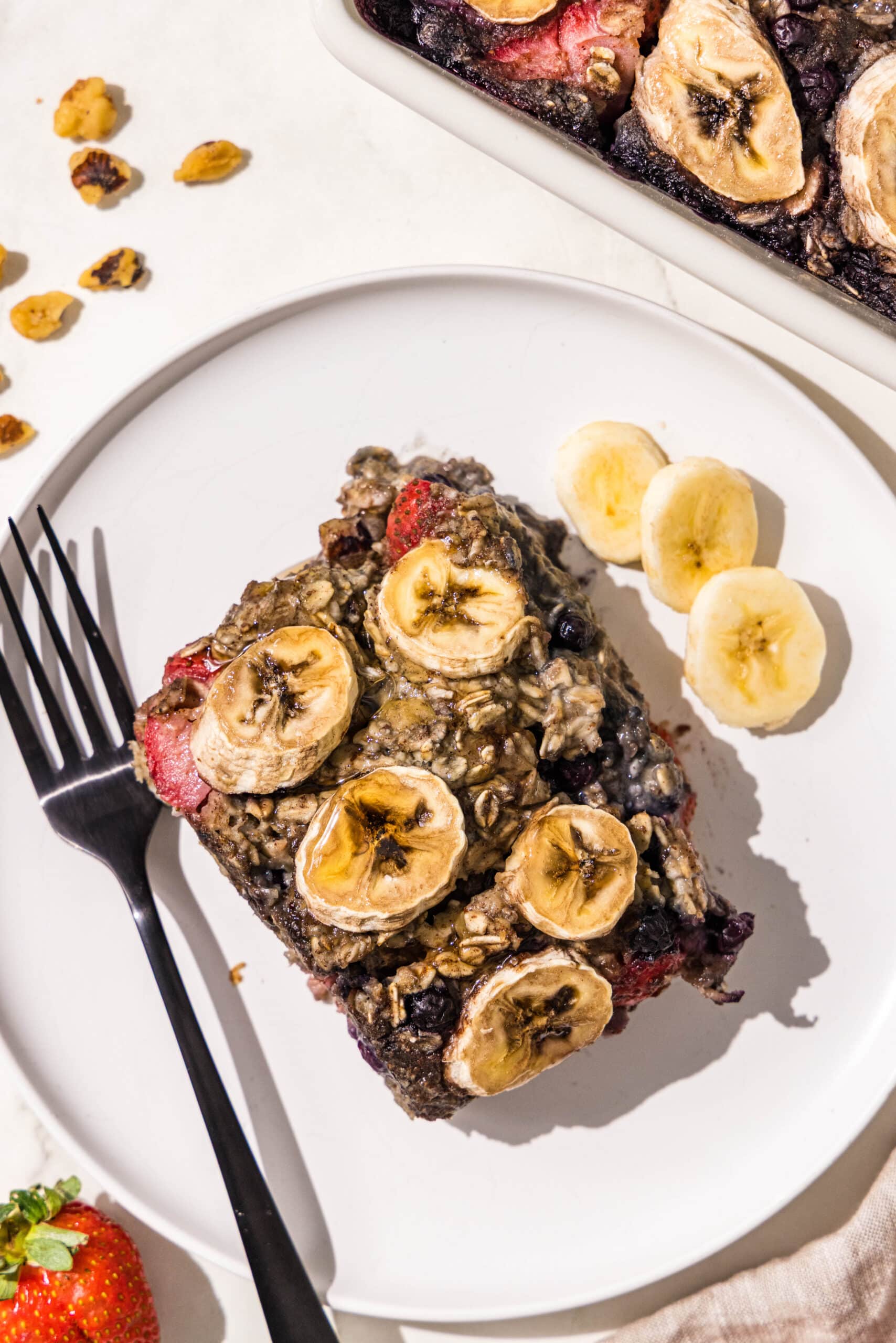 A piece of baked oatmeal topped with bananas on a white plate with a black fork and a couple slices of banana.