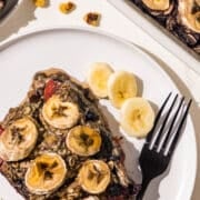 A piece of baked oatmeal topped with bananas on a white plate with a black fork and a couple slices of banana and 1 strawberry.