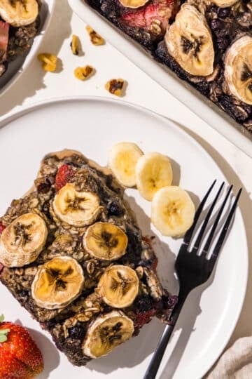 A piece of baked oatmeal topped with bananas on a white plate with a black fork and a couple slices of banana and 1 strawberry.
