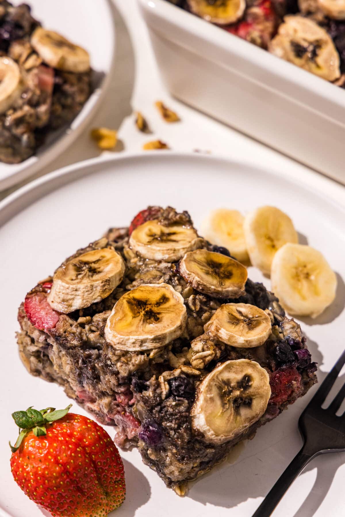 A piece of baked oatmeal topped with banana slices and a strawberry on a white plate with a black fork.