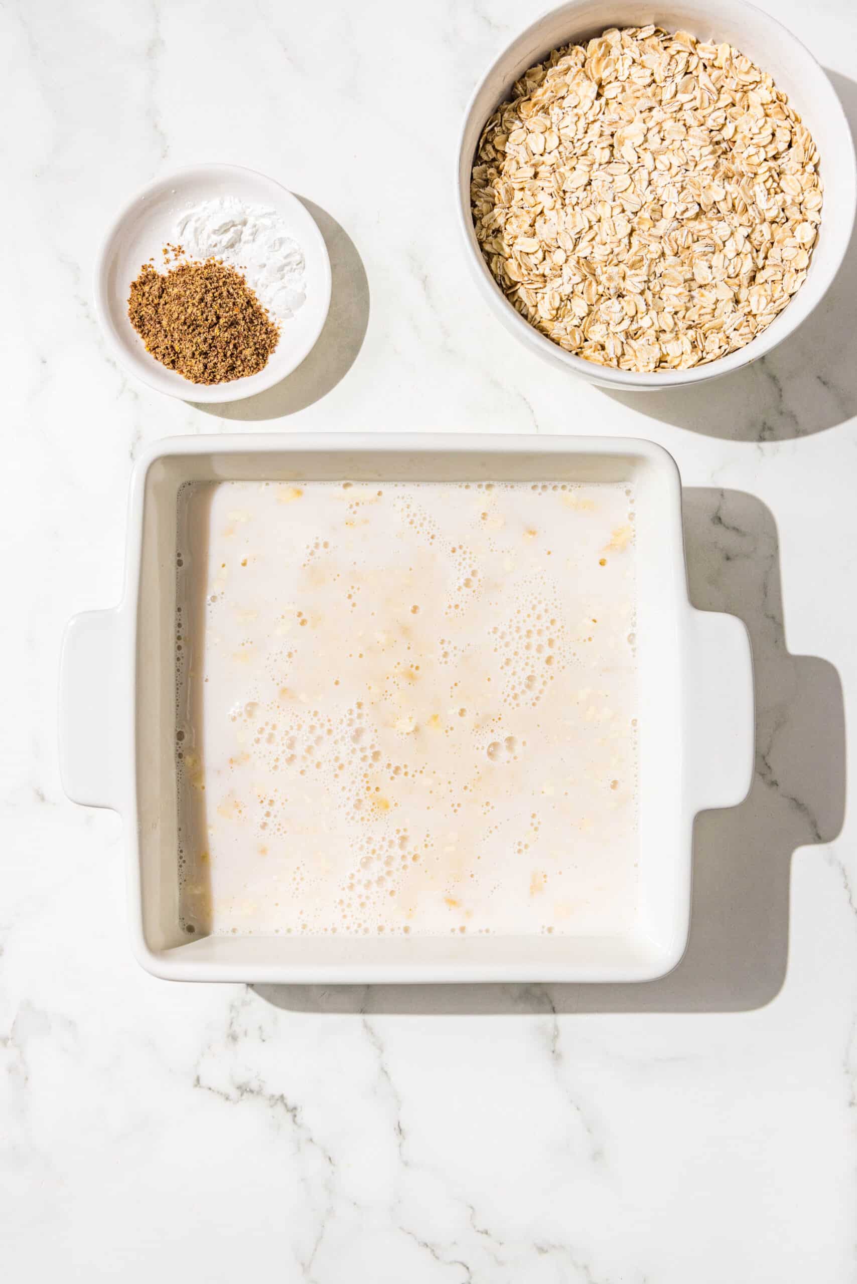 A 9x9 baking dish filled with almond milk and mashed bananas. Beside the dish is a bowl of oats and a bowl of flax seed.
