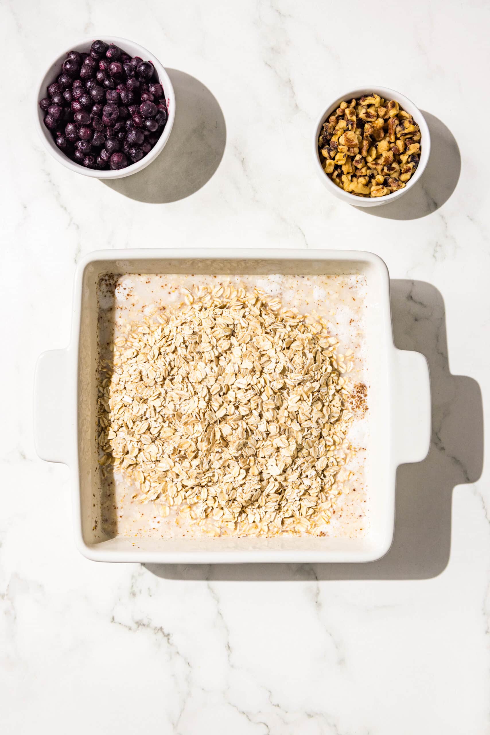 A 9x9 baking dish filled with almond milk, mashed bananas, and oats. Beside the dish is two small bowls of frozen blueberries and walnuts.