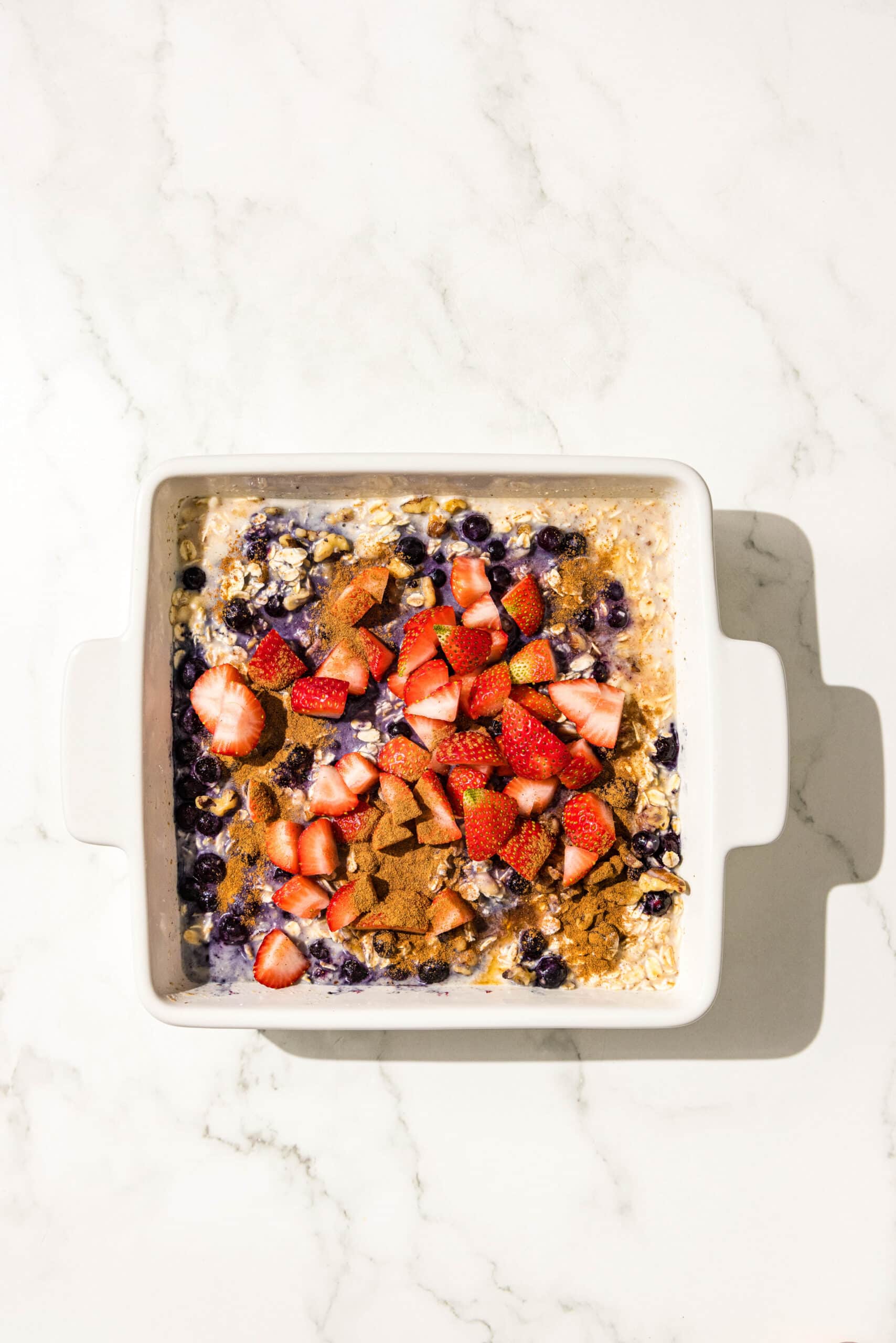 A 9x9 baking dish filled with oats, almond milk, mashed banana, wild blueberries, chopped strawberries, walnuts, and cinnamon.
