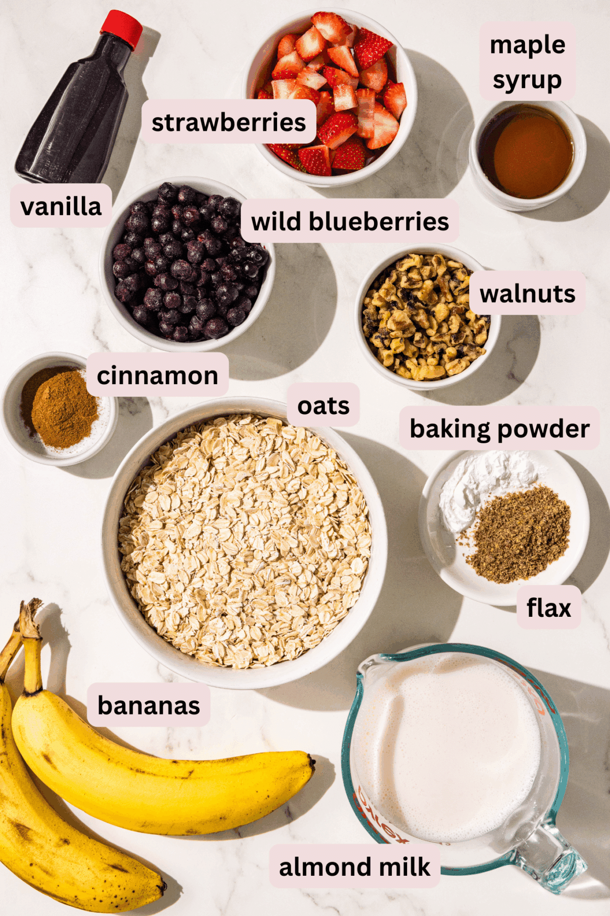 Ingredients laid out on a counter in bowls and labled including: vanilla extract, strawberries, maple syrup, wild blueberries, walnuts, oats, cinnamon, baking powder, flax, bananas, almond milk.
