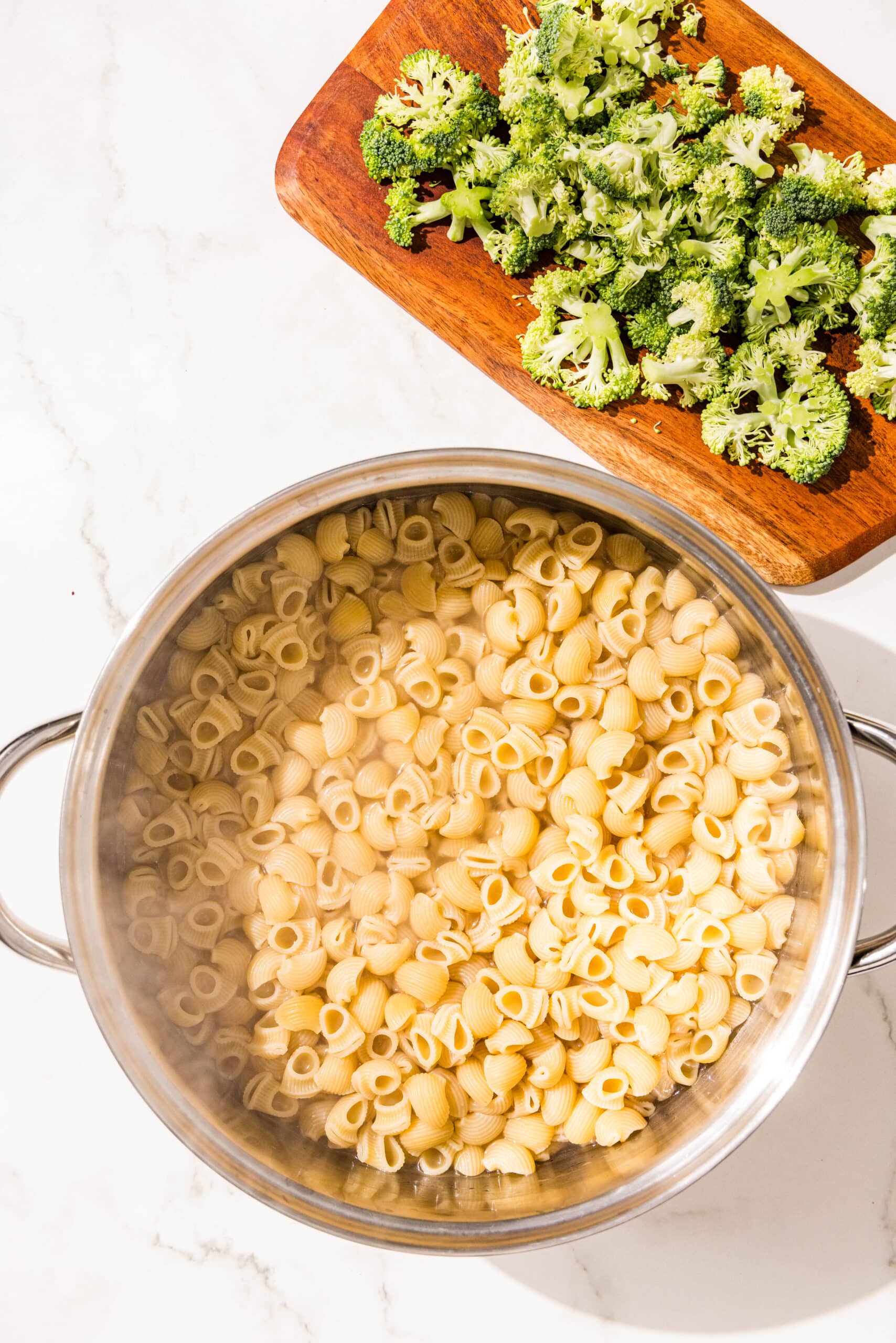 A silver pot of cooked pasta and a small cutting board with broccoli on it.