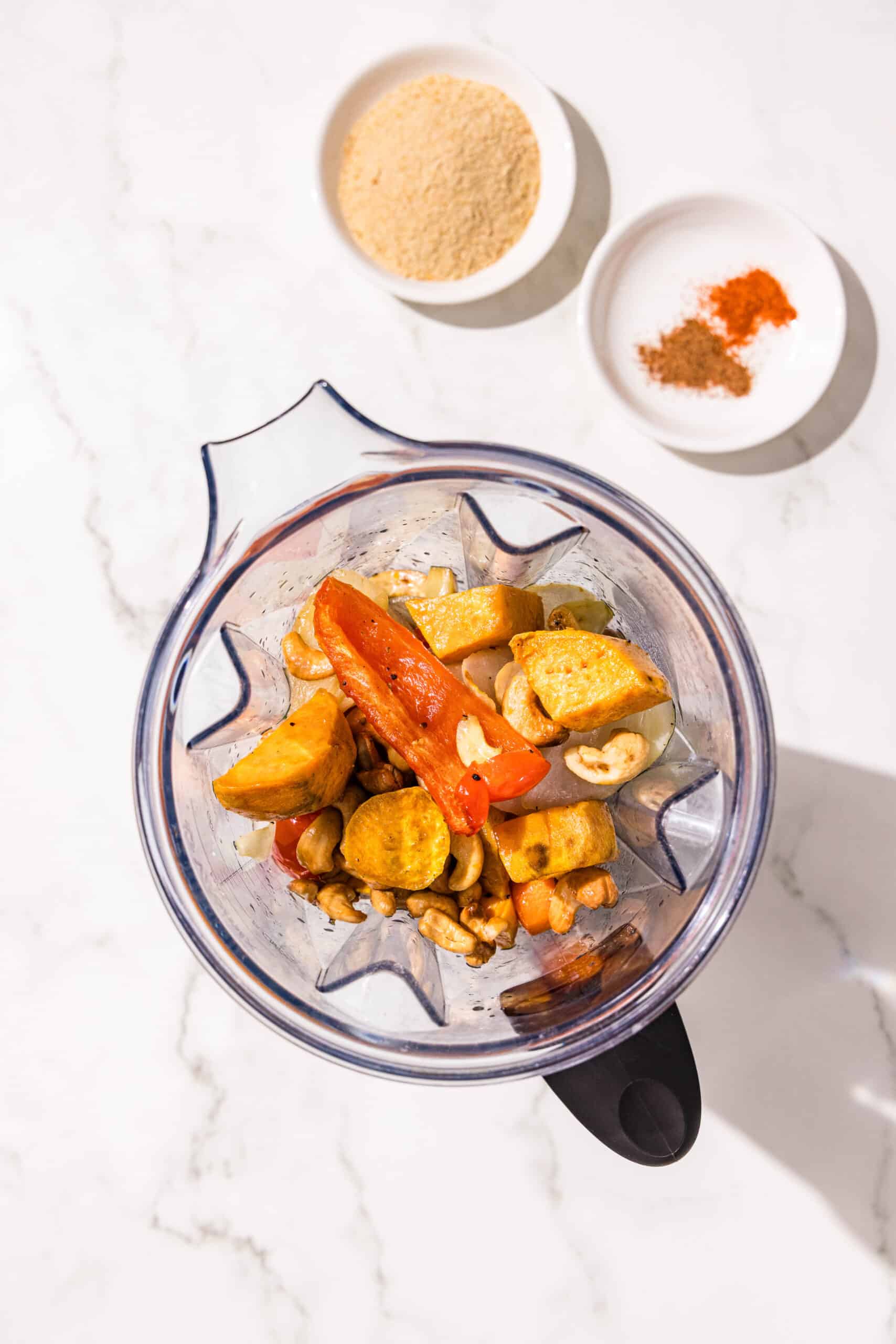 A blender filled with roasted veggies and two small bowls of spices and nutritional yeast.