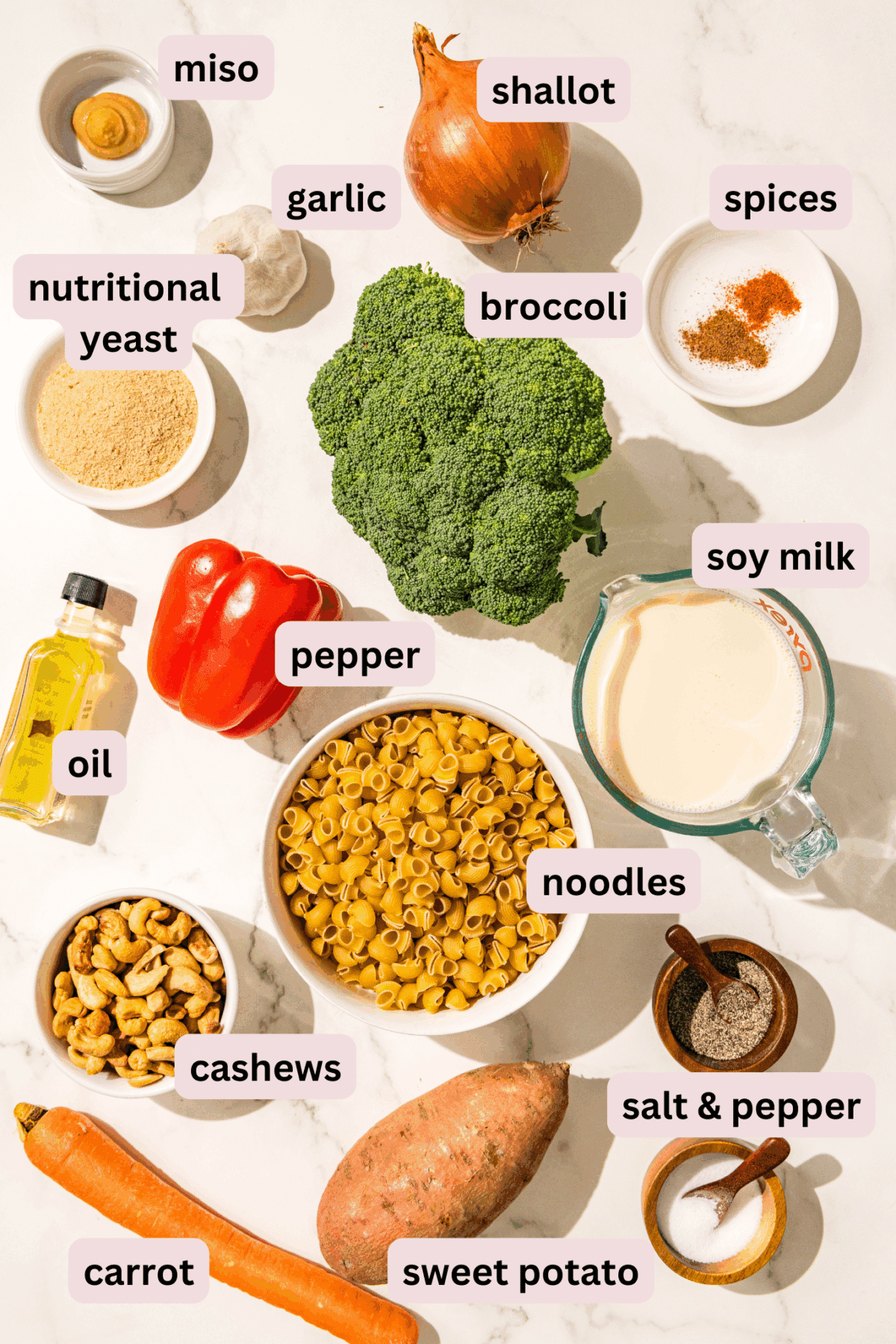 Ingredients laid out on the counter in bowls and labeled including: miso, shallot, spices, garlic, broccoli, nutritional yeast, soy milk, bell pepper, oil, noodles, salt and pepper, cashews, carrot, and sweet potato.